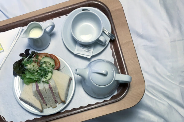 Photograph looking down at a tray containing an empty cup and saucer, a small teapot, a jug of milk and a plate with triangular ham sandwiches and salad to the left