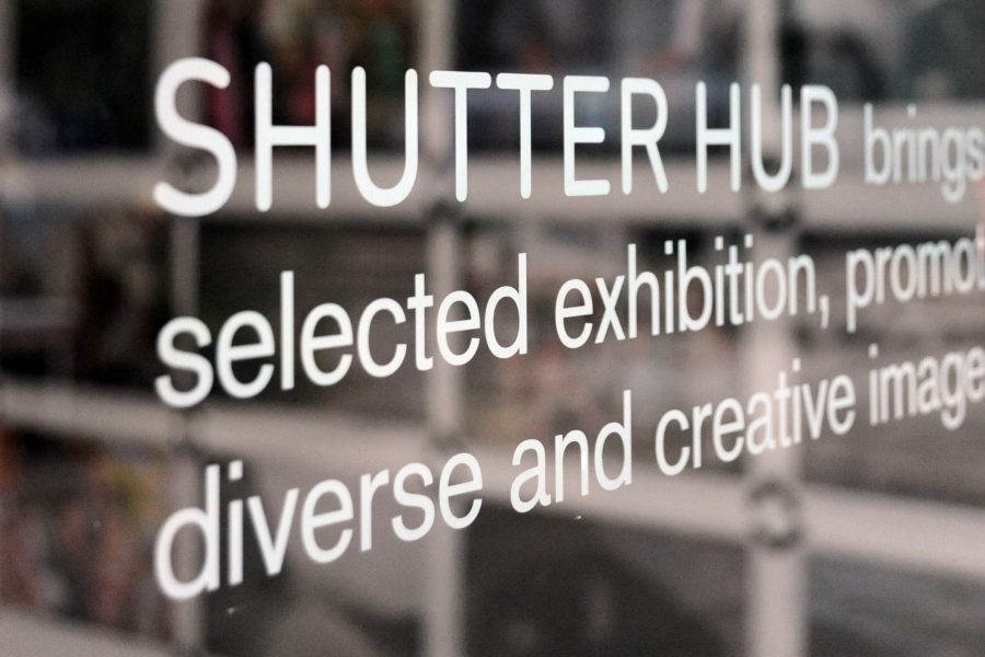 Photograph of white text on a window, with photographs from an exhibition out of focus in the background. The text says 'SHUTTER HUB brings, selected exhibition, promoting, diverse and creative images'