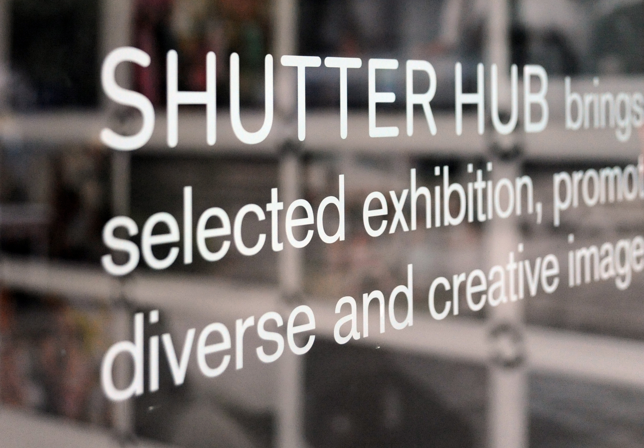 Photograph of white text on a window, with photographs from an exhibition out of focus in the background. The text says 'SHUTTER HUB brings, selected exhibition, promoting, diverse and creative images'