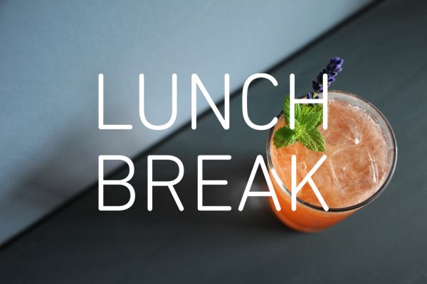 Photograph of an orange-coloured, fruit drink viewed from above, with ice, a sprig of mint and lavender. Photographed from above the table and the wall make a diagonal line with light and dark grey section. The words 'LUNCH BREAK' are in the centre in white text.