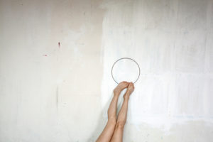 Photograph of legs against a painted white wall holding a large ring with their feet