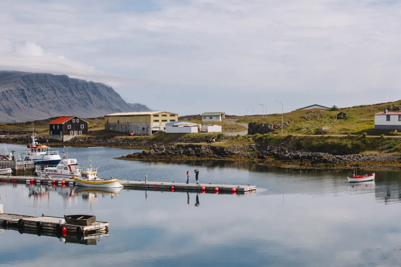Fishing boats and a quiet dock in Djúpivogur harbor with low buildings and mountains behind.