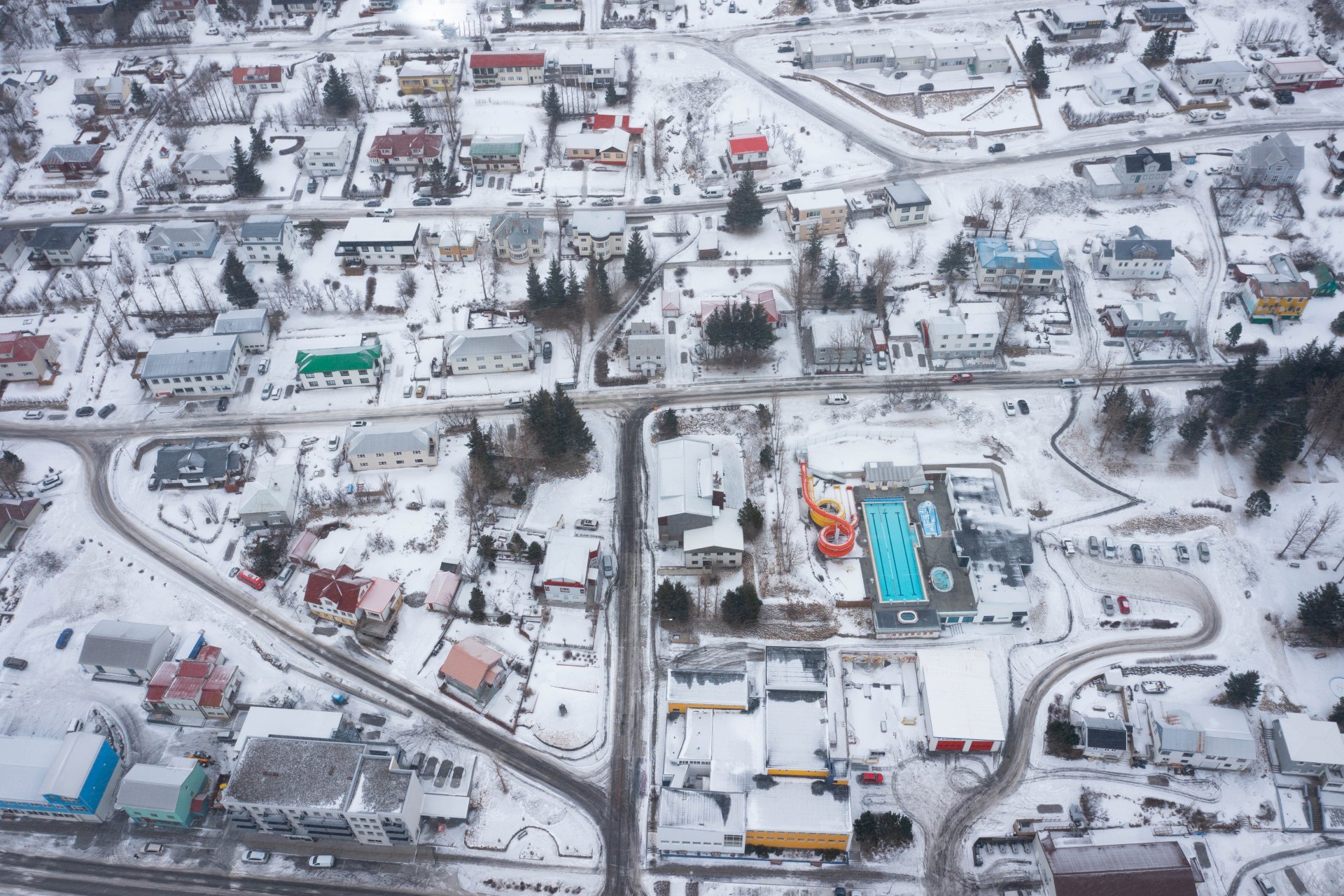 Winter aerial over an East Iceland town with snowy streets, low buildings, and compact neighborhood blocks.
