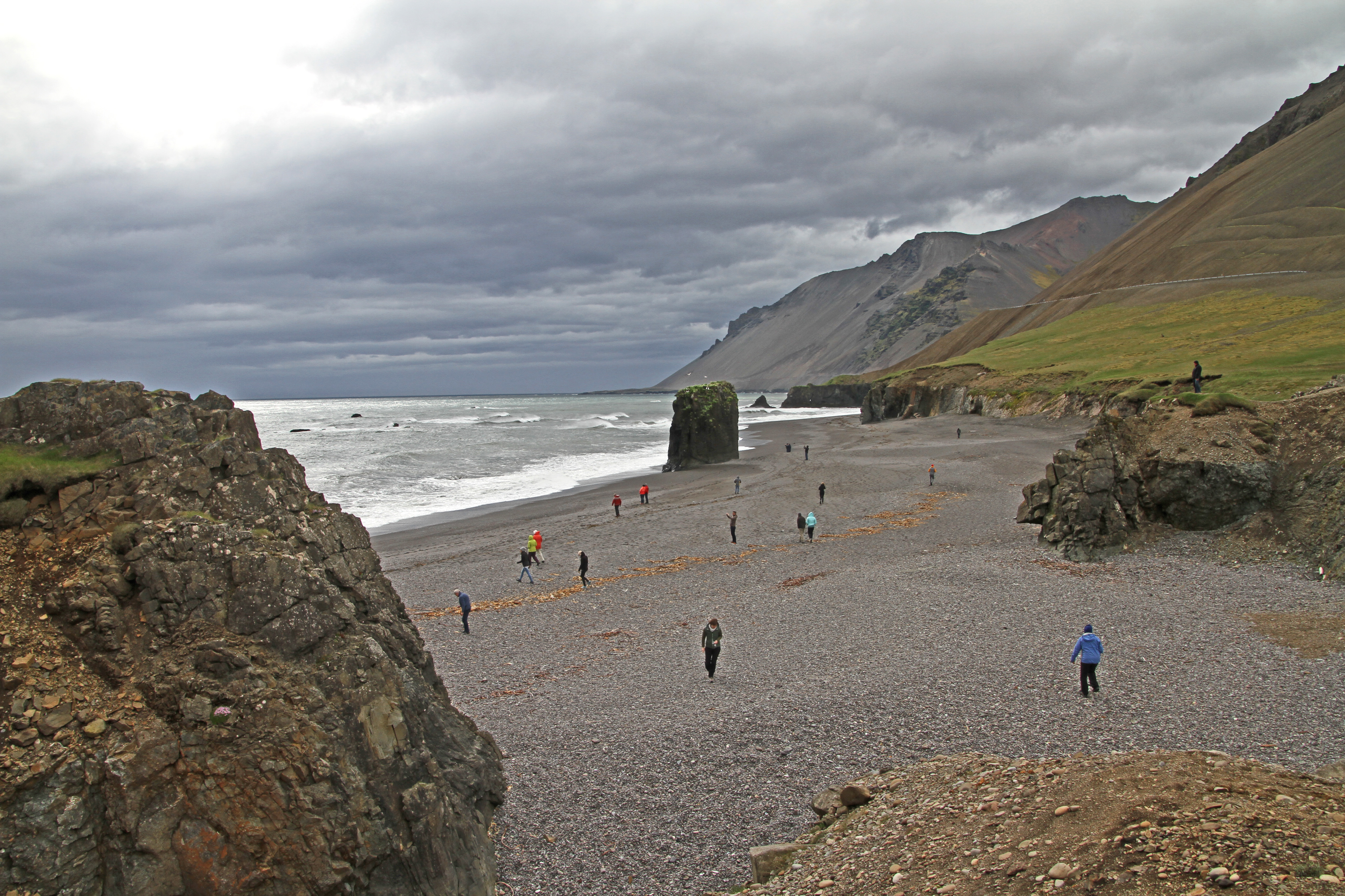 Fauskasandur viewed from above, with the dark beach, sea stack, surf, and steep Hvalnes mountain wall behind it.