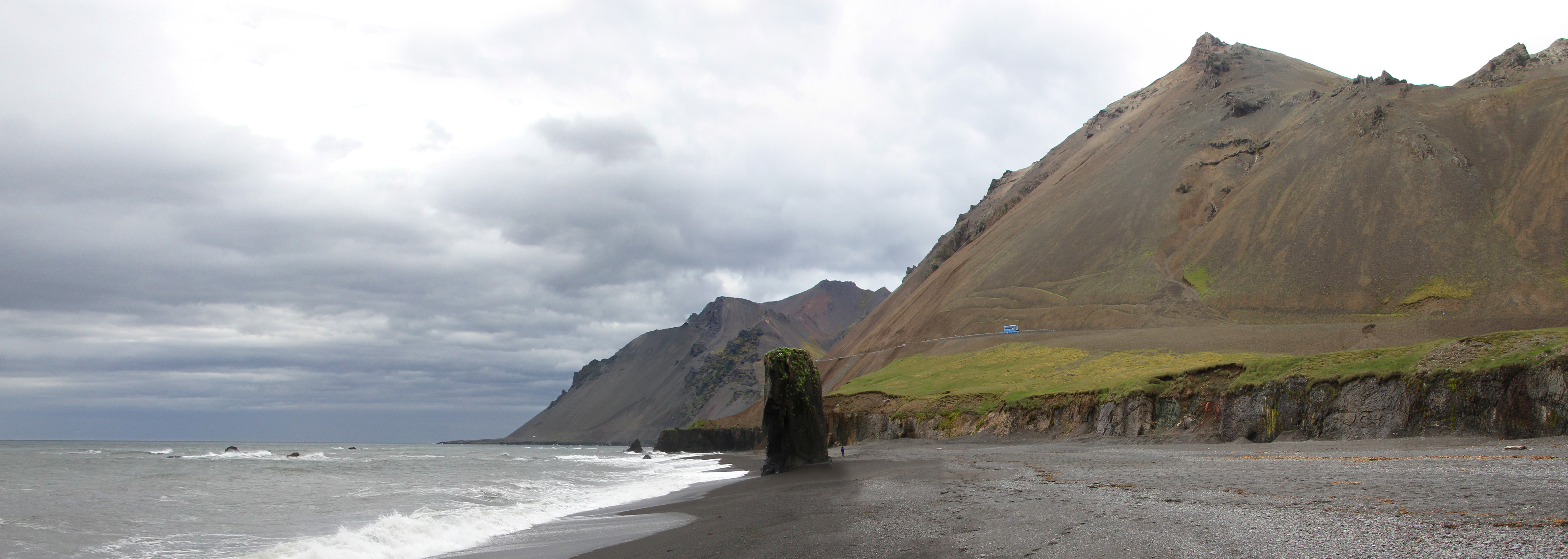 Panoramic view across Fauskasandur with the sea stack, beach curve, and roadside mountain slope near Hvalnes.