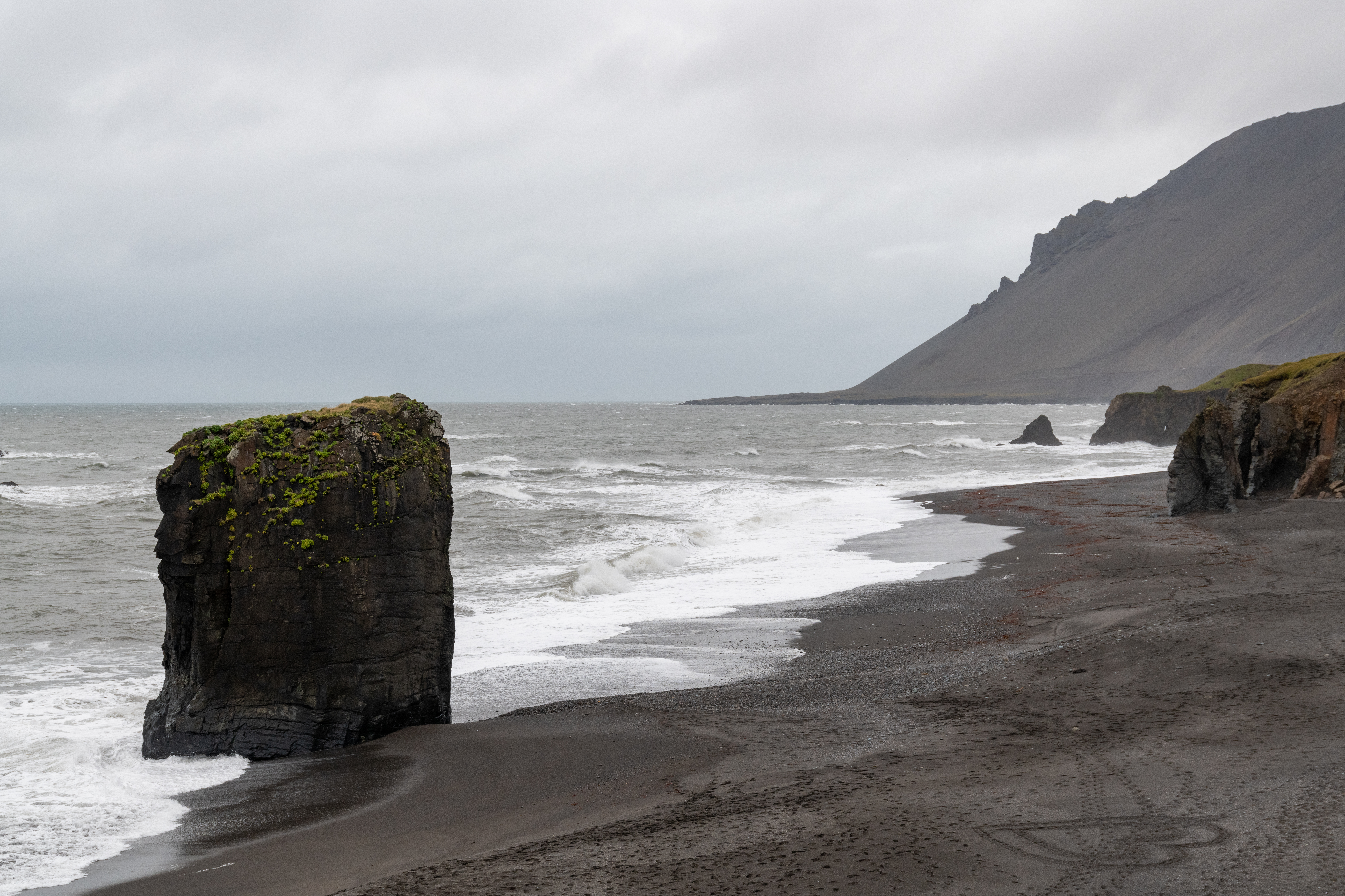 Close view of the Fauskasandur sea stack beside dark sand and white surf under the Hvalnes mountain slope.