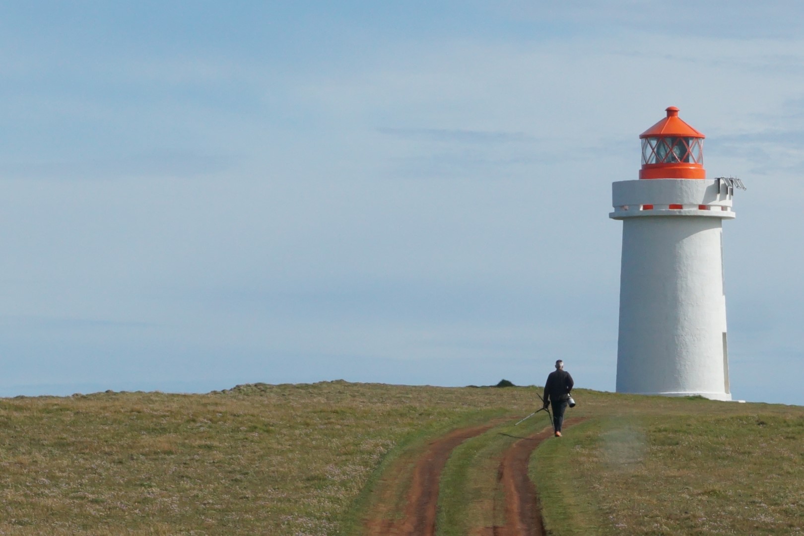 A person walks toward the white Fontur lighthouse along a grassy vehicle track.