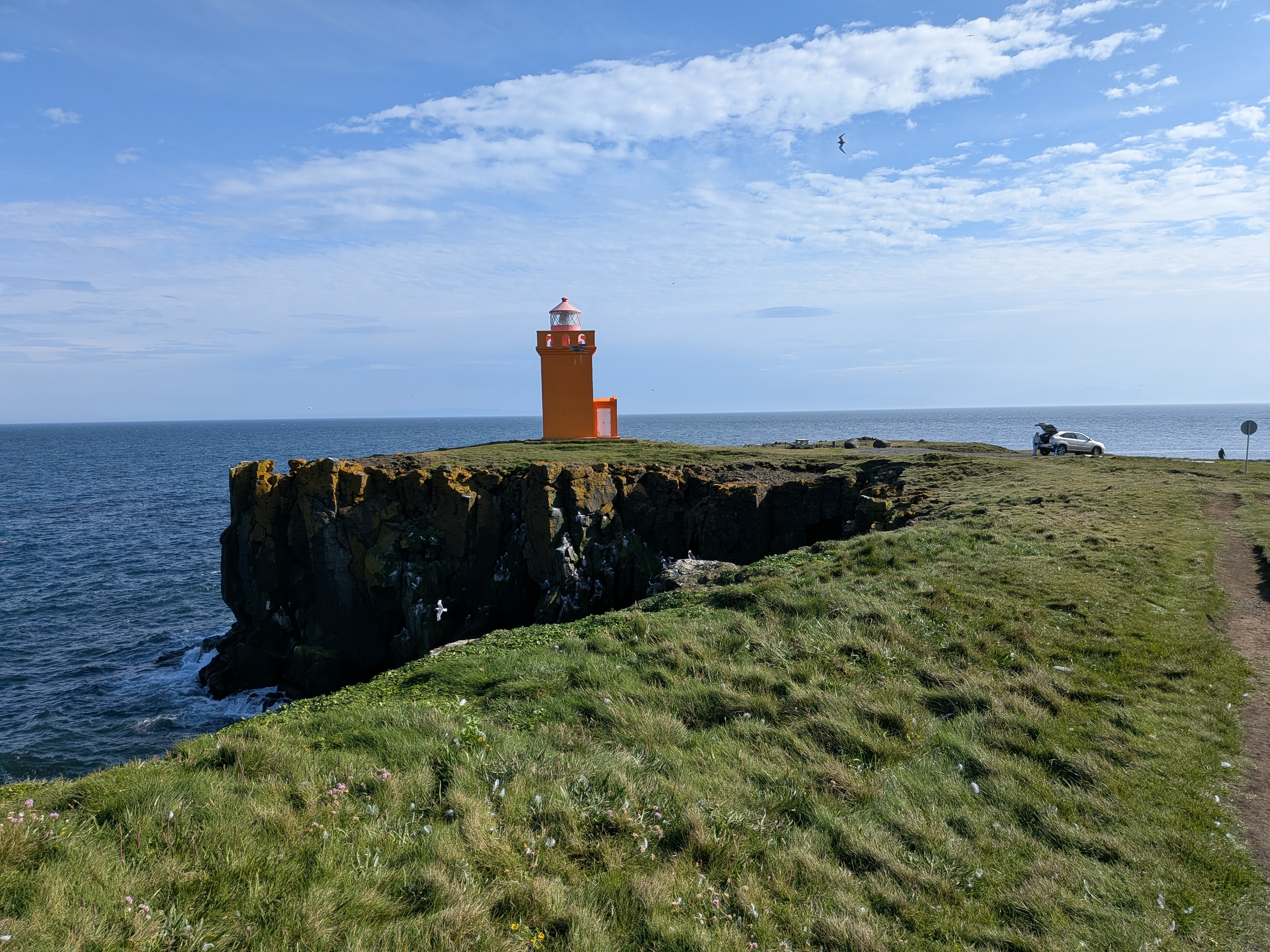 White Grímsey lighthouse on grassy coastal ground with the sea behind it.