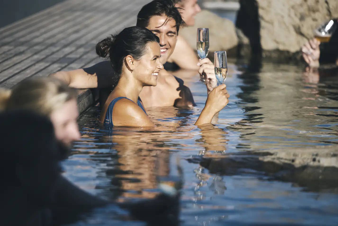 Visitor with a drink in the water at Hvammsvik Hot Springs with the fjord behind.