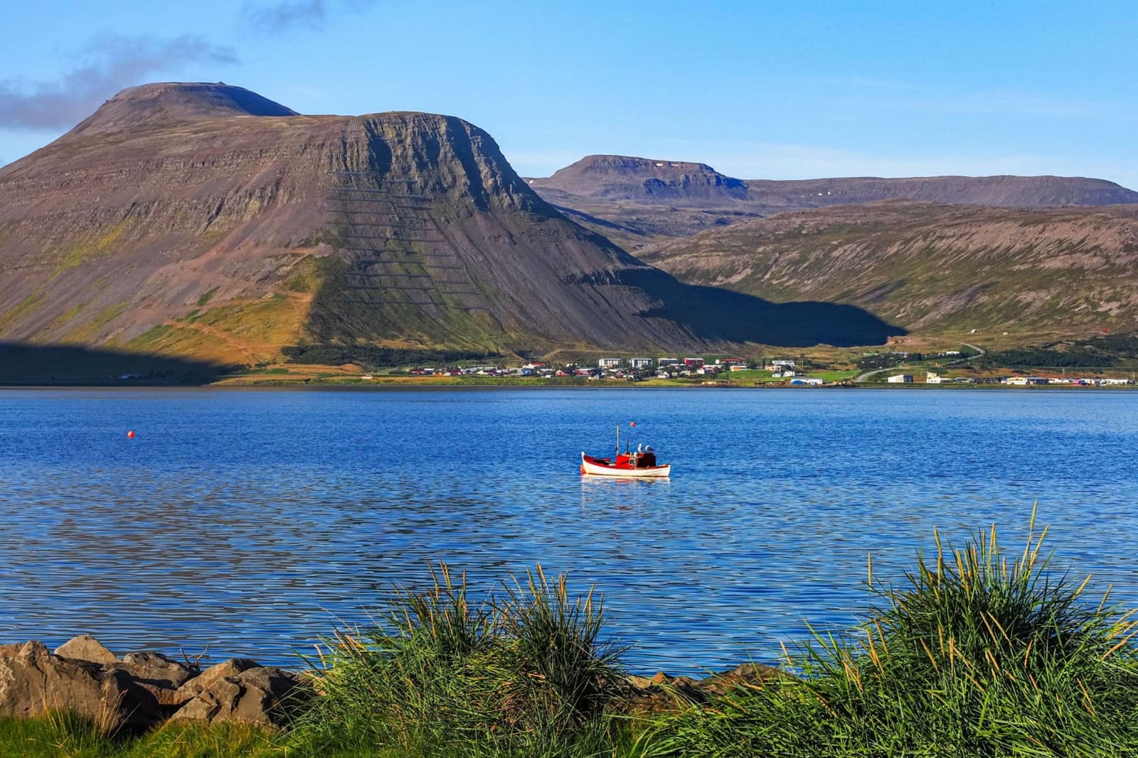Fishing boat on Ísafjörður fjord with town buildings and Westfjords mountains reflected in calm water.