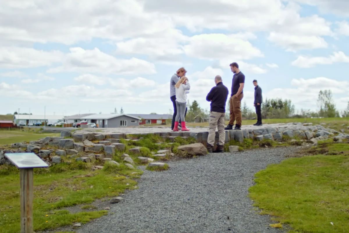 Visitors beside the marked approach to Kirkjugólf with the basalt edge, gravel path, and village buildings nearby.