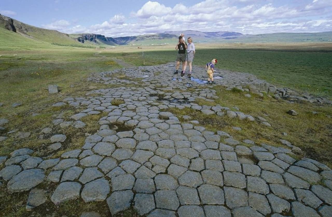 Visitors standing on Kirkjugólf's basalt pavement with open South Iceland fields and distant mountains behind them.