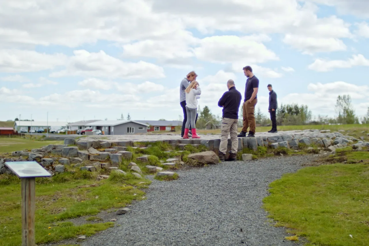 Visitors beside the marked approach to Kirkjugólf with the basalt edge, gravel path, and village buildings nearby.