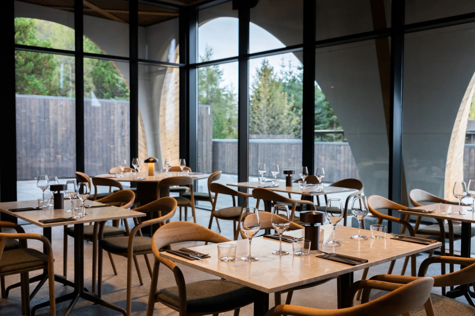 Tables and curved wooden chairs beside tall windows in Ylja Restaurant at Laugarás Lagoon.