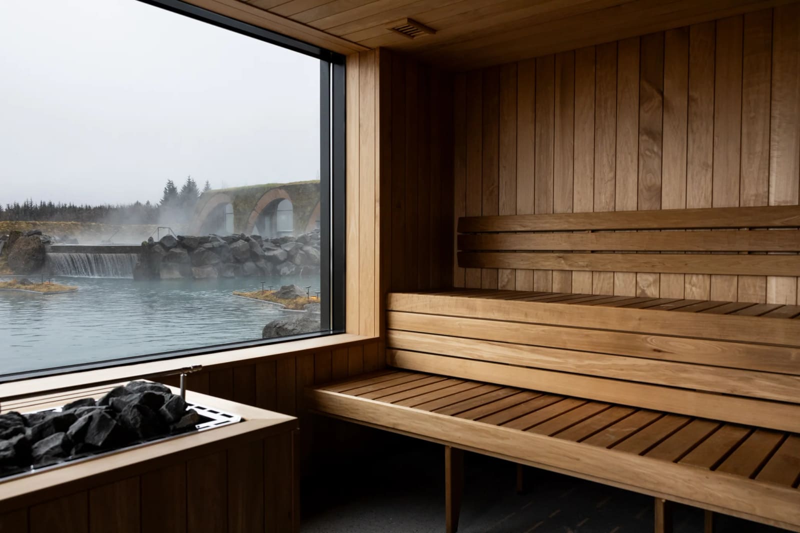 Timber benches and a stone heater inside the sauna at Laugarás Lagoon, with a window over the water.