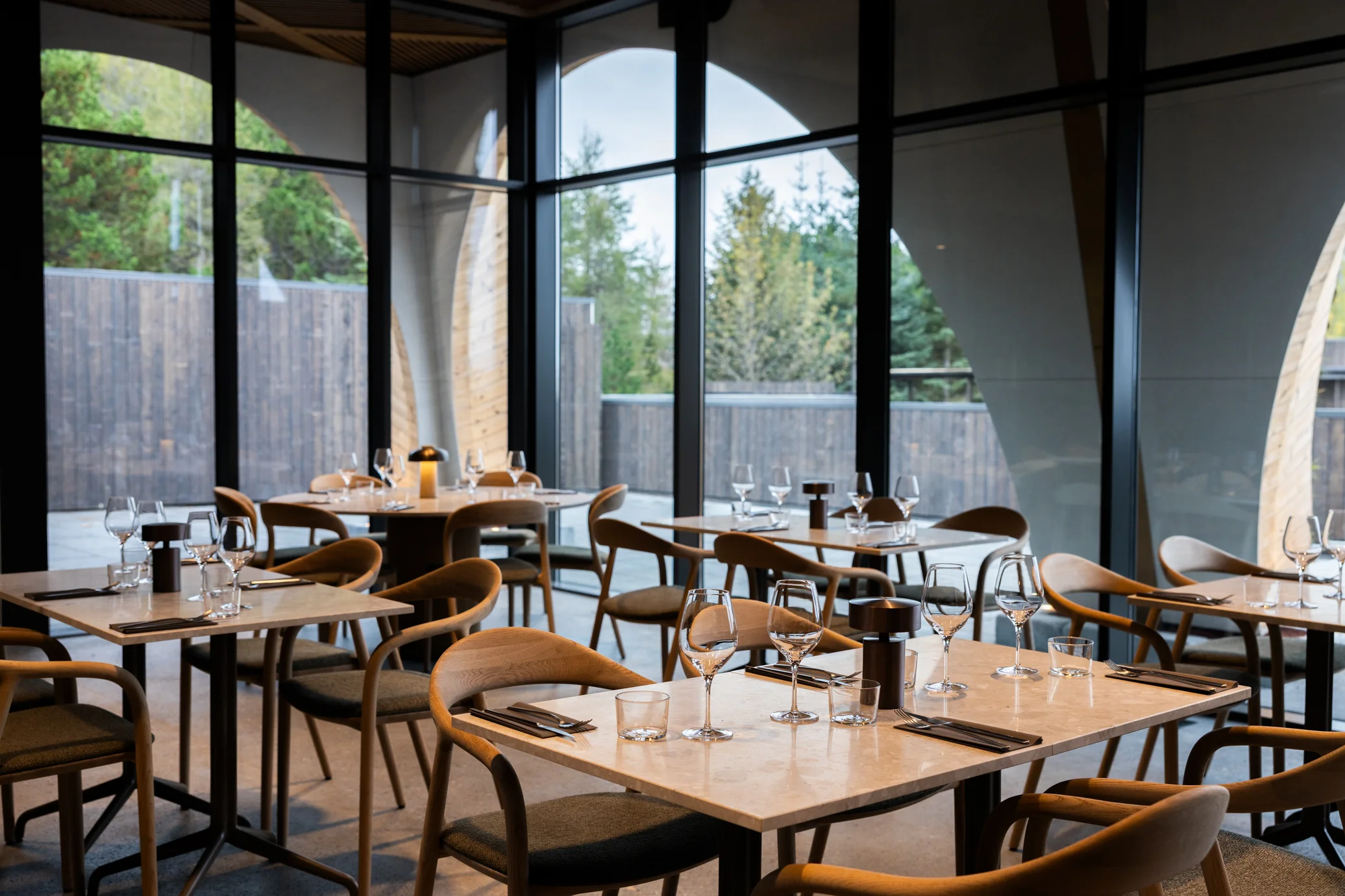 Tables and curved wooden chairs beside tall windows in Ylja Restaurant at Laugarás Lagoon.