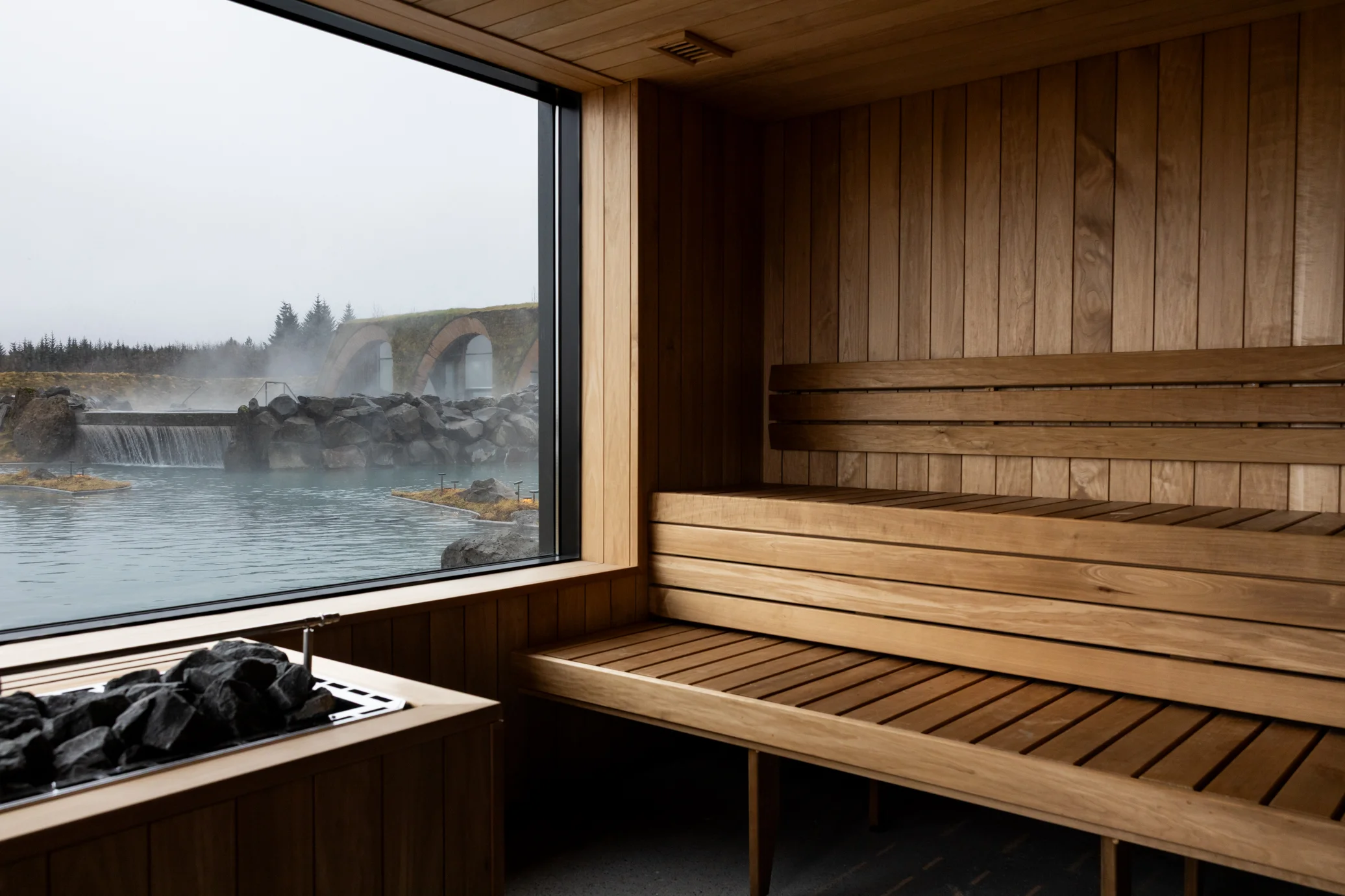 Timber benches and a stone heater inside the sauna at Laugarás Lagoon, with a window over the water.