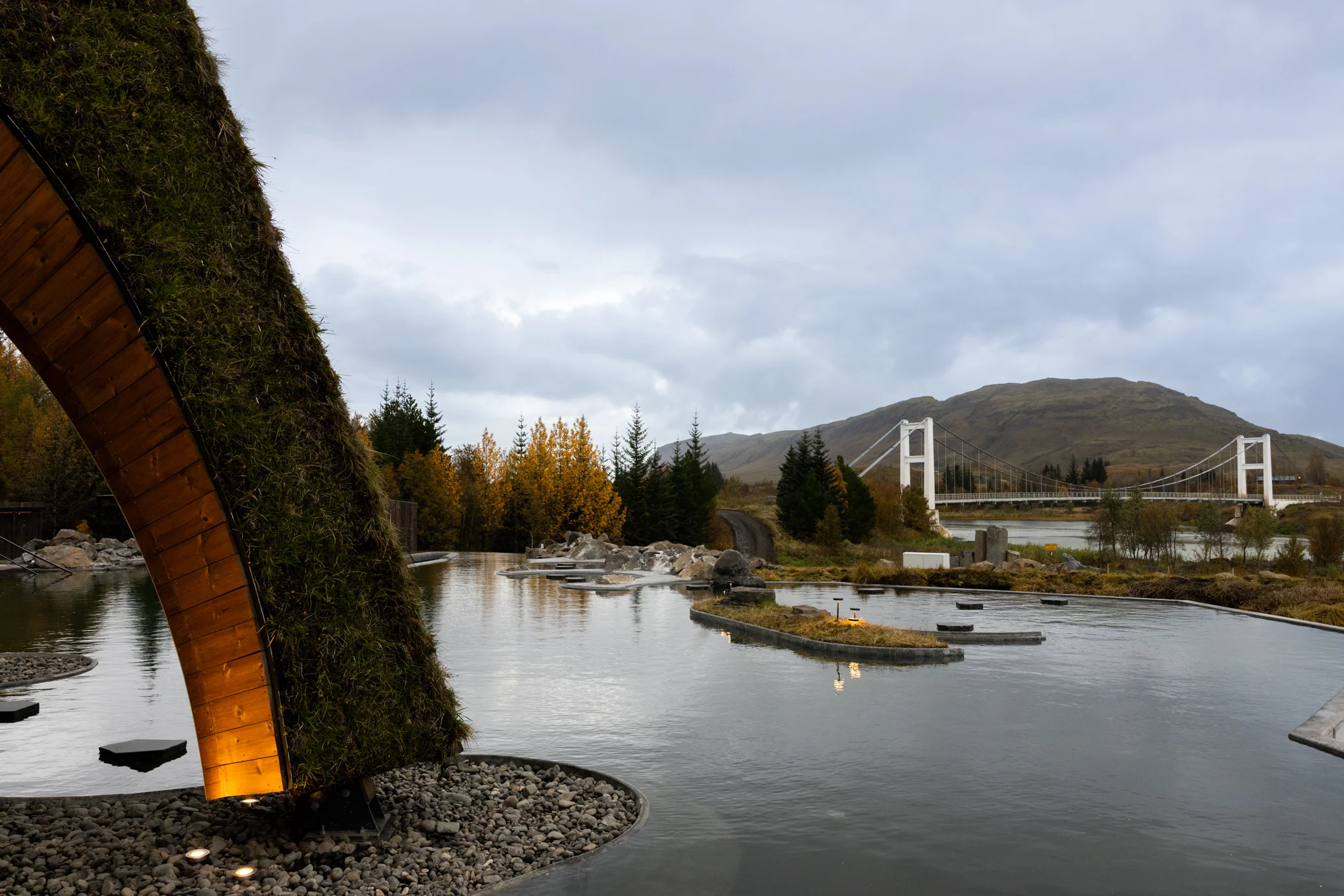 Laugarás Lagoon beneath a turf-roofed arch, with the Hvítá bridge and a hill beyond.
