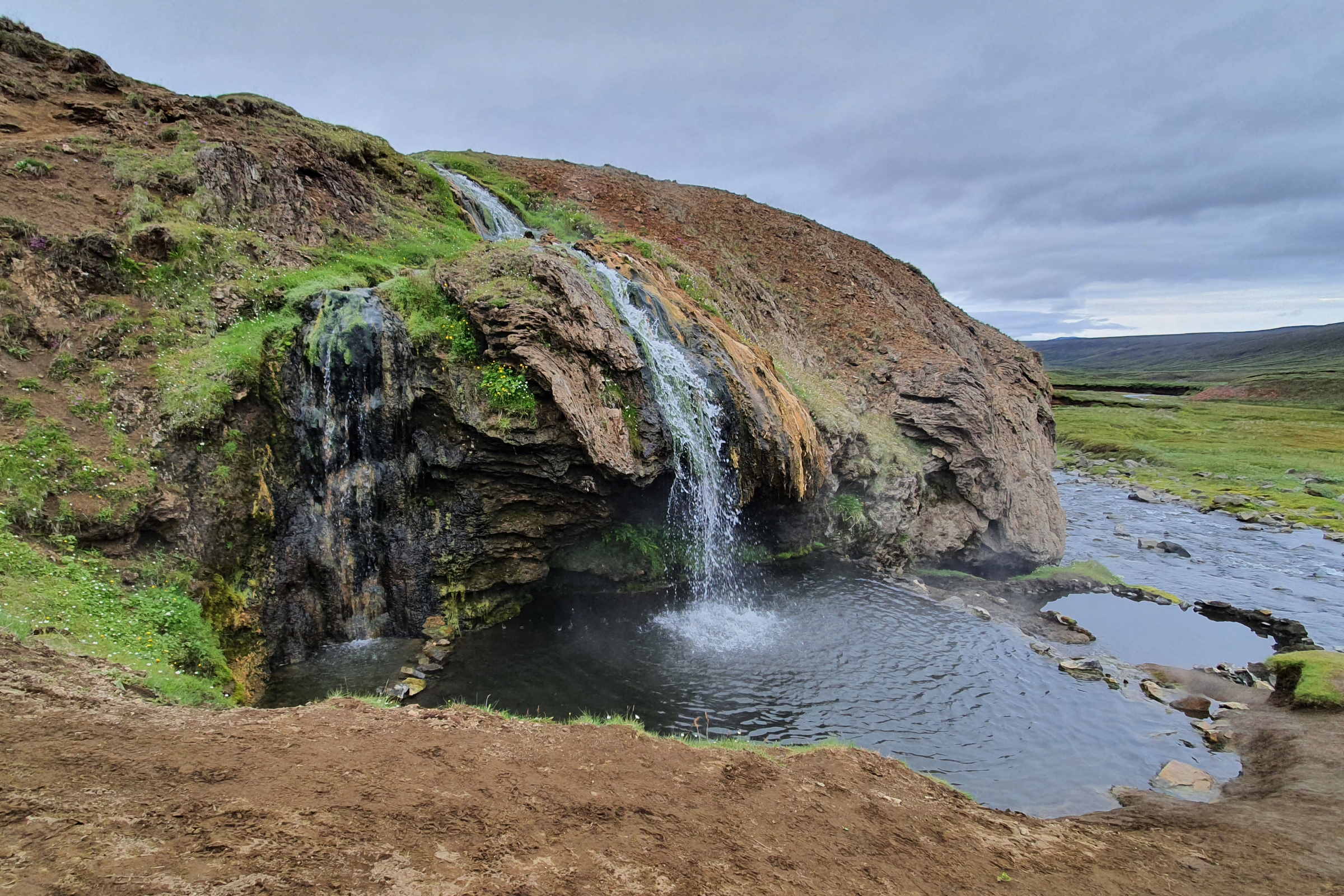 Laugavallalaug hot spring pool below a small warm waterfall in a green highland valley.