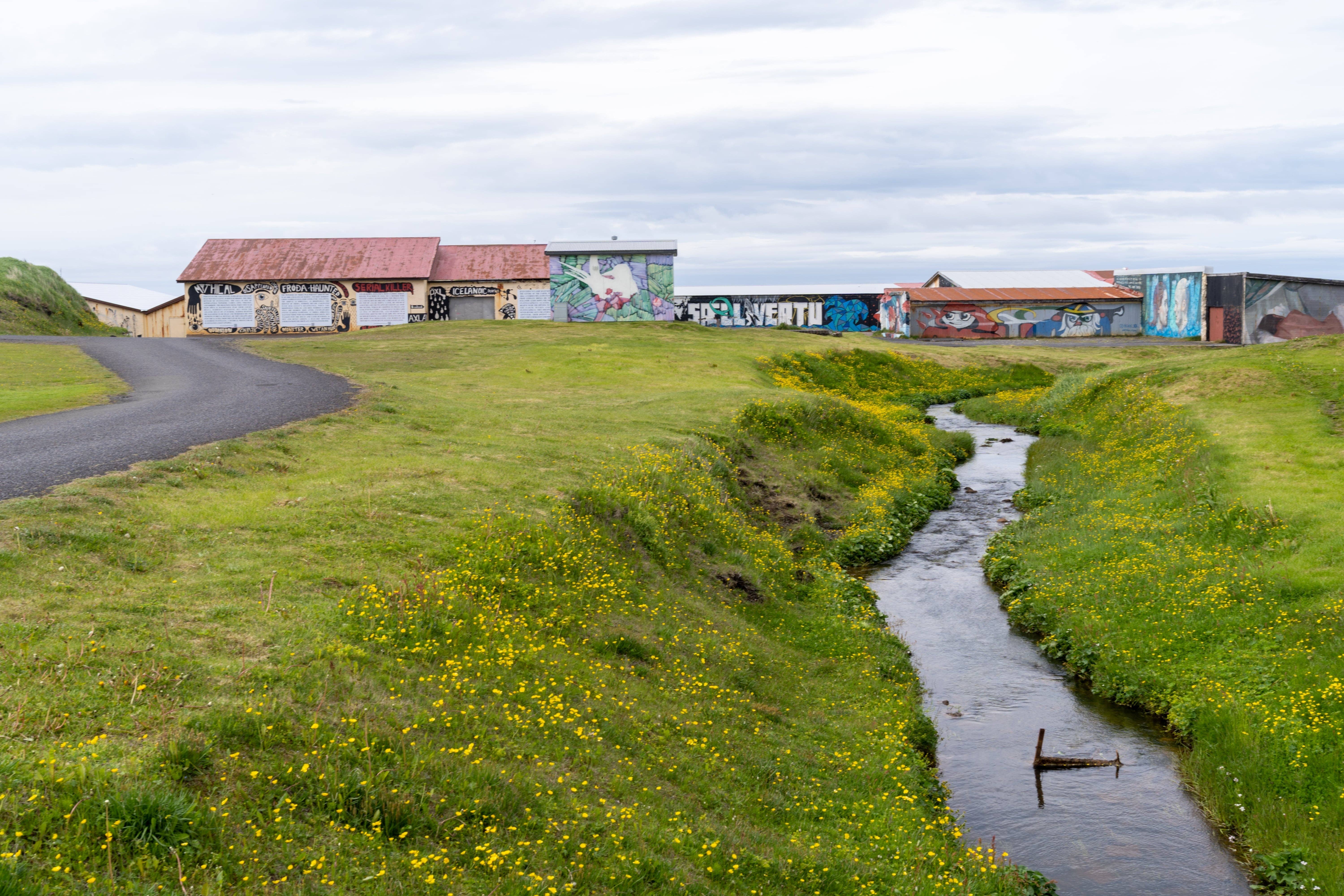 Murals on low buildings beside a stream and grassy banks in Hellissandur.