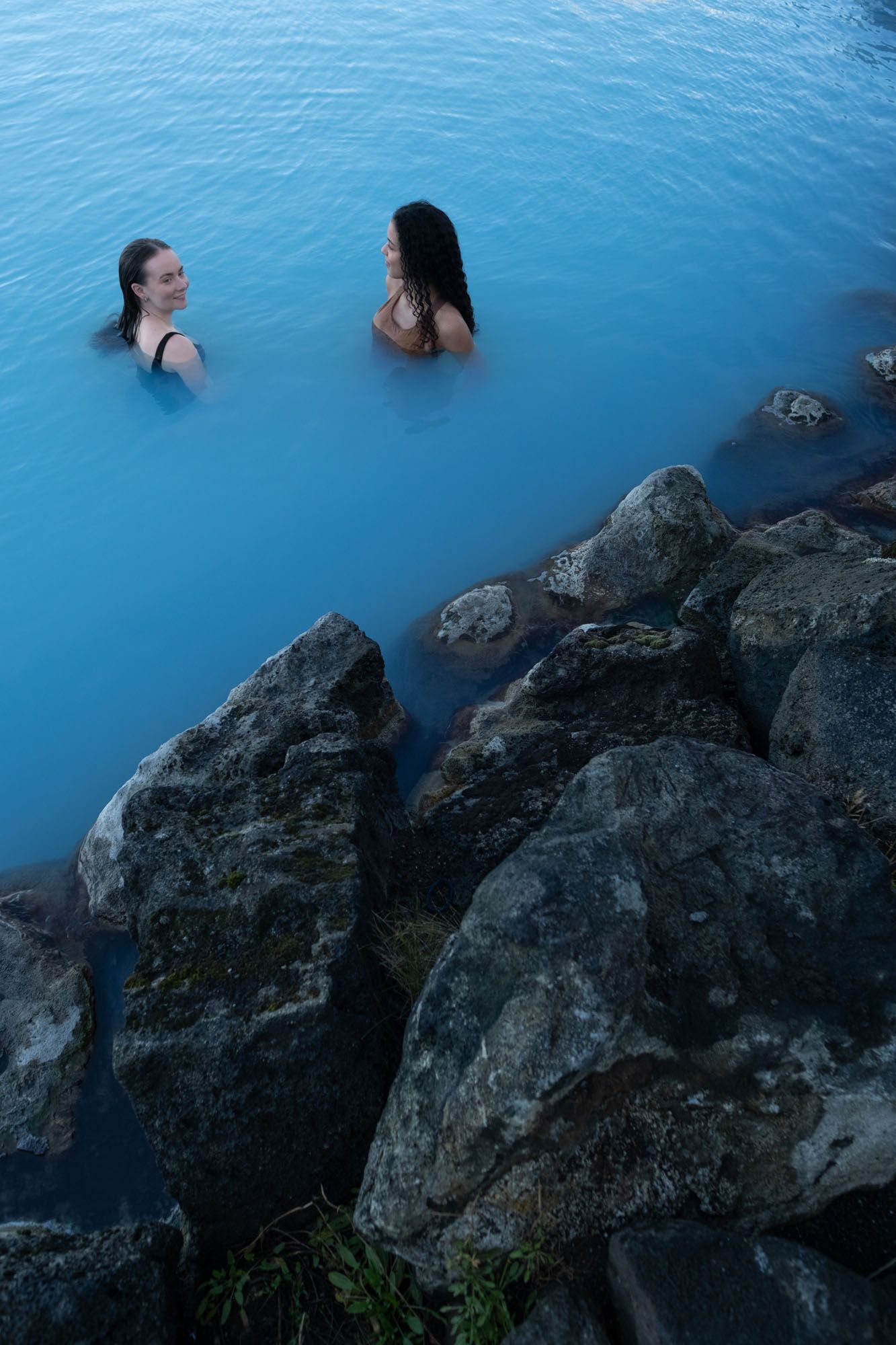 Visitor standing in turquoise water beside the dark stone exterior of Earth Lagoon Mývatn.