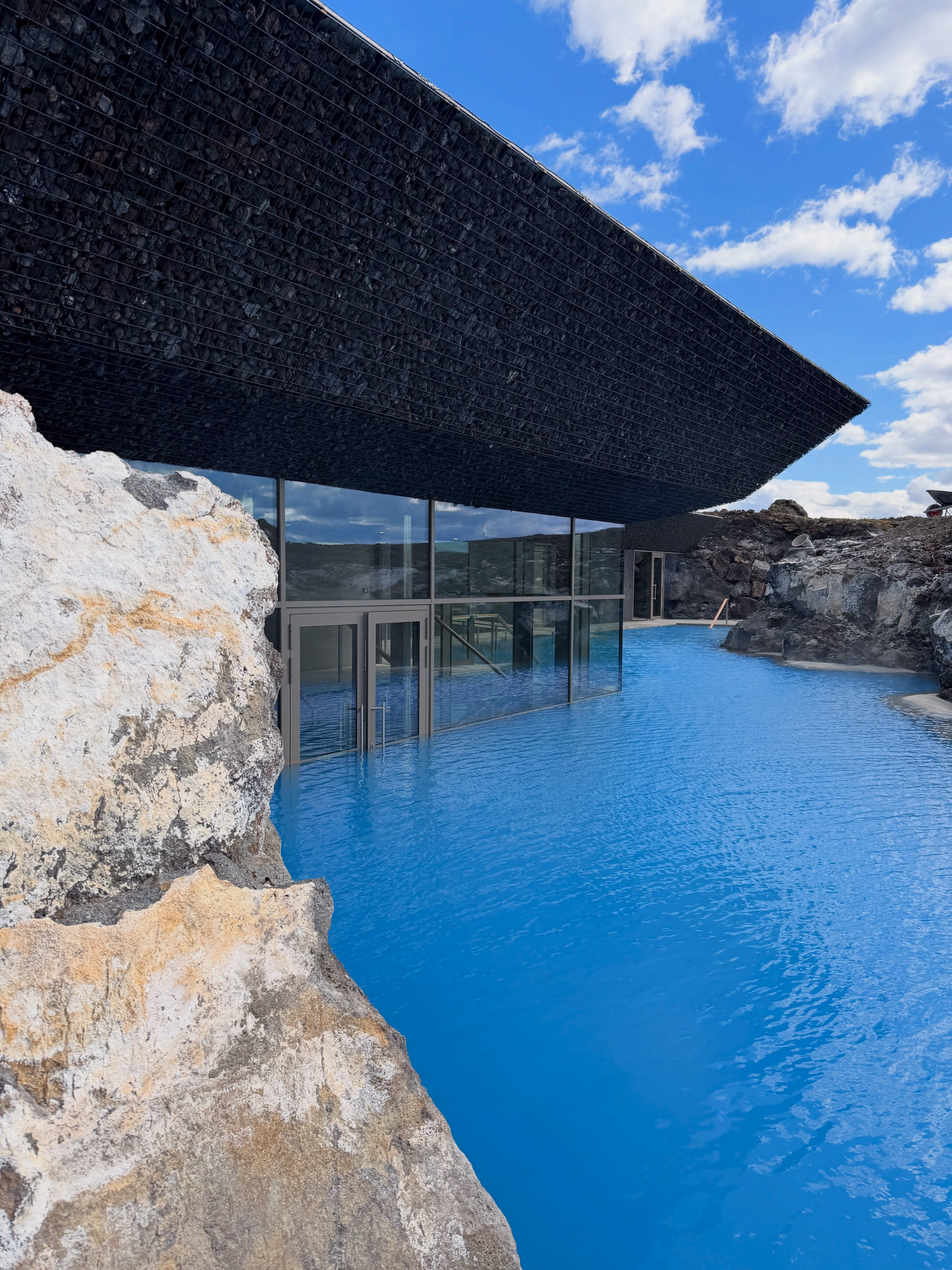 Glass entrance and dark stone canopy meeting the expanded blue lagoon at the rebuilt Earth Lagoon Mývatn.
