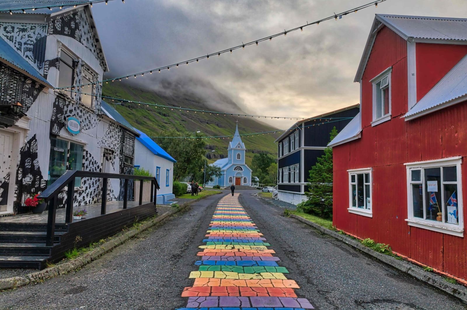 The Blue Church at the end of Seyðisfjörður’s Rainbow Street with colorful town buildings and steep fjord mountains behind it.
