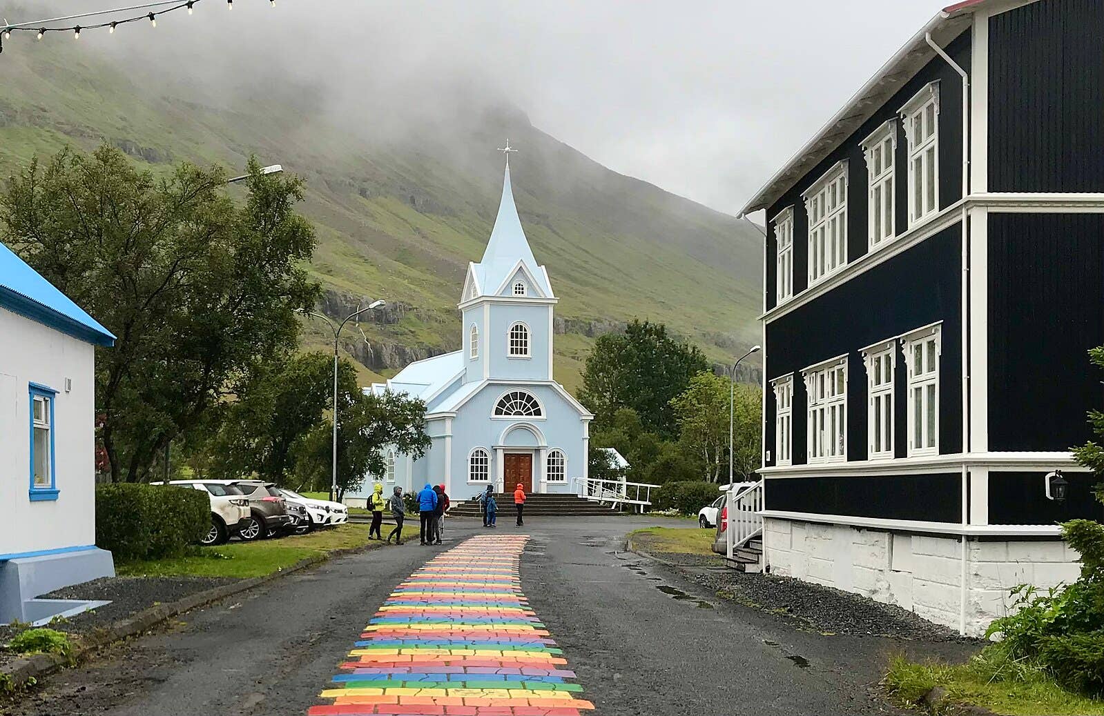 Rainbow Street leading toward Seyðisfjörður Church with visitors, parked cars, and low clouds on the mountain behind.
