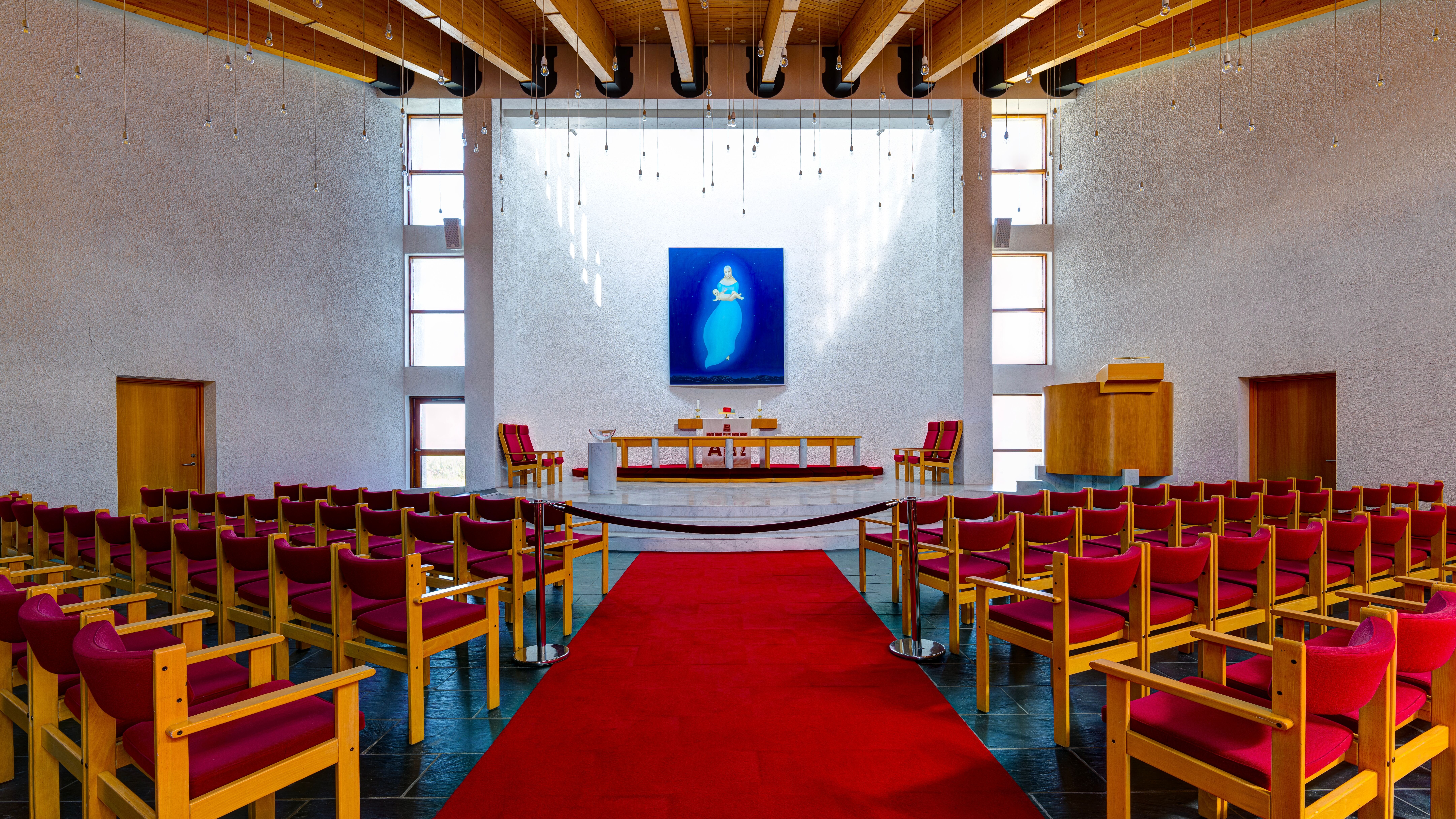 Interior of Stykkishólmskirkja Church with red chairs, altar, hanging lights, and tall white walls.