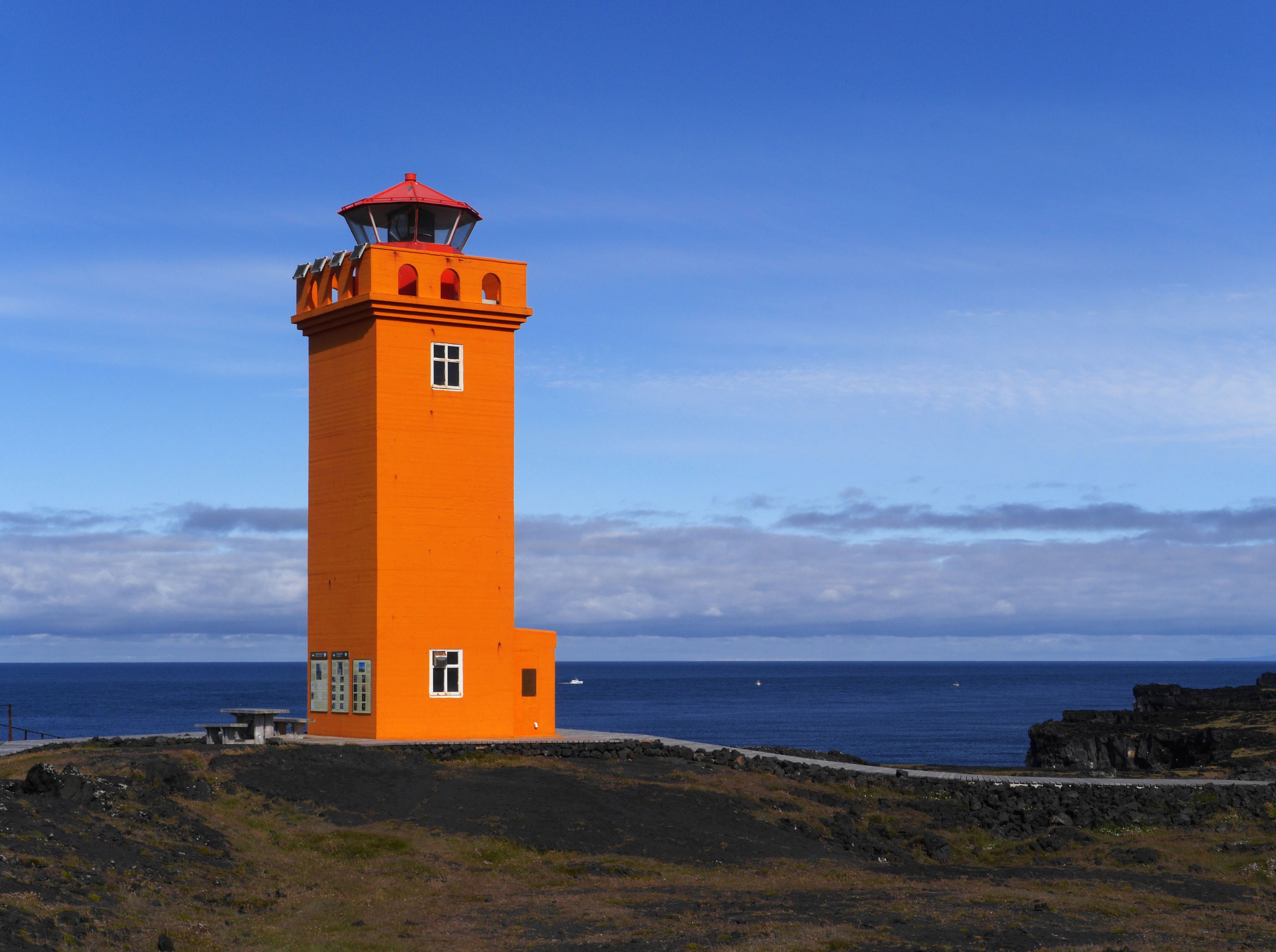 Close view of the orange Svörtuloft lighthouse above the dark lava ground and ocean edge.