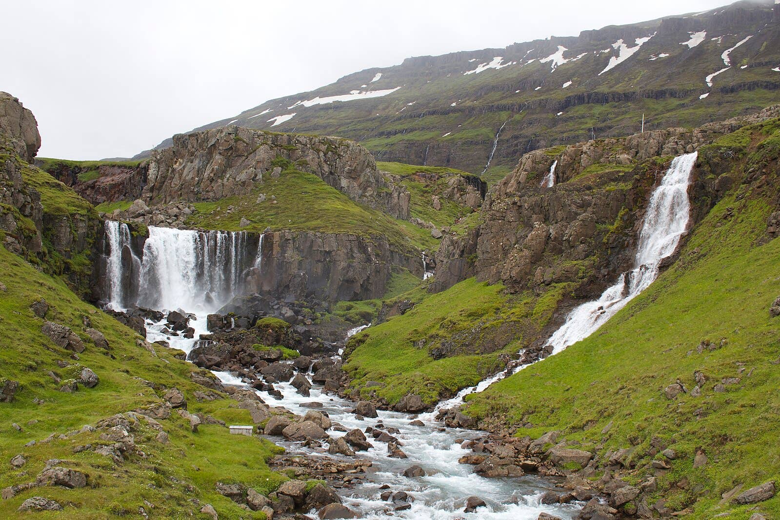 Several Vestdalsfossar waterfalls descending through a rocky green valley near Seyðisfjörður.
