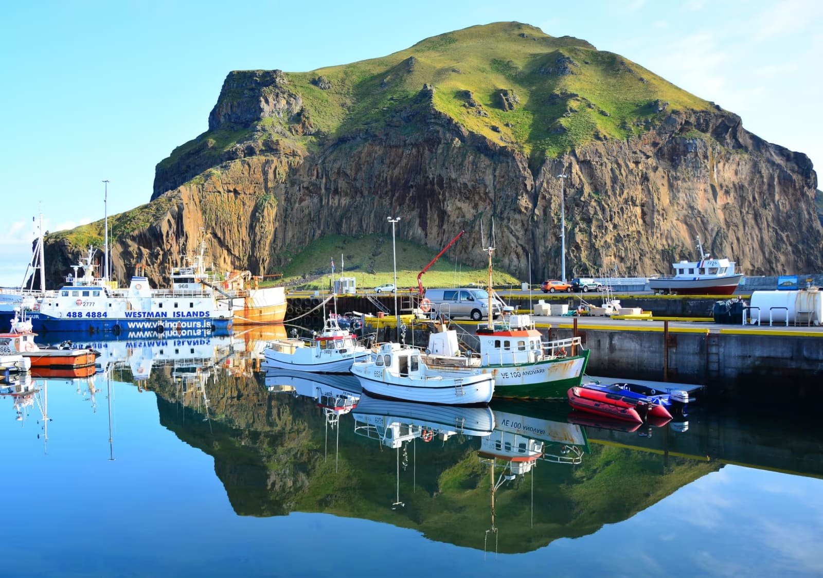 Fishing boats reflected in Heimaey harbor below steep cliffs in the Westman Islands.
