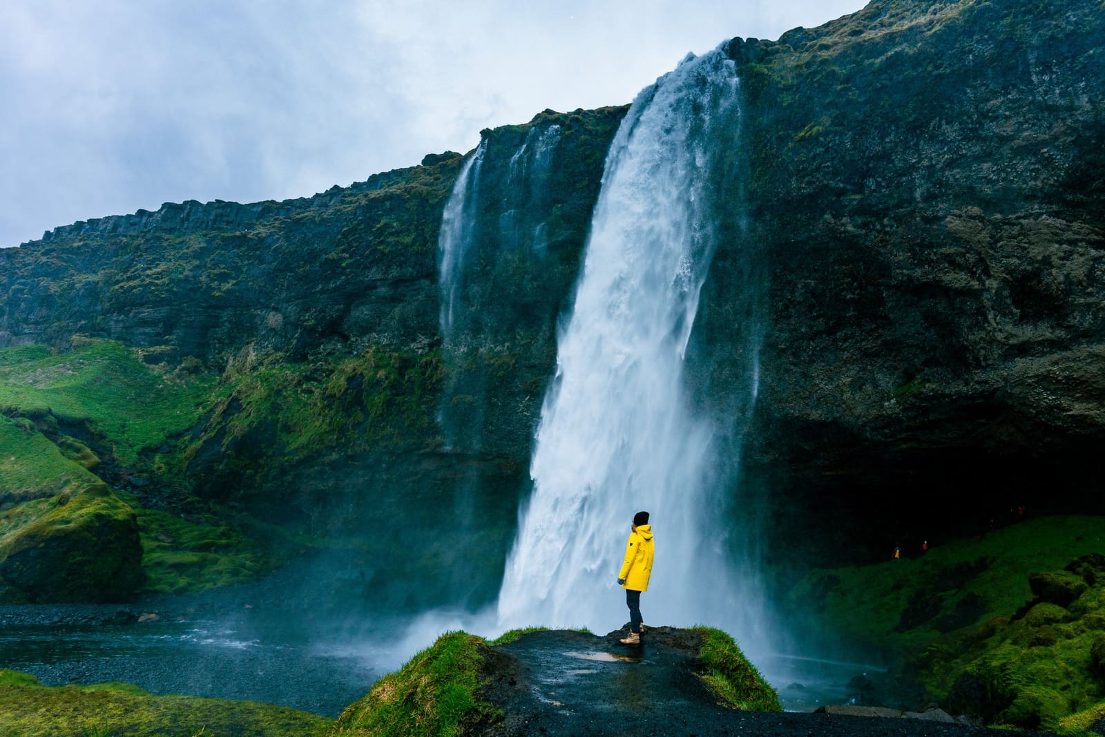 Visitor in a yellow coat standing beside Seljalandsfoss waterfall