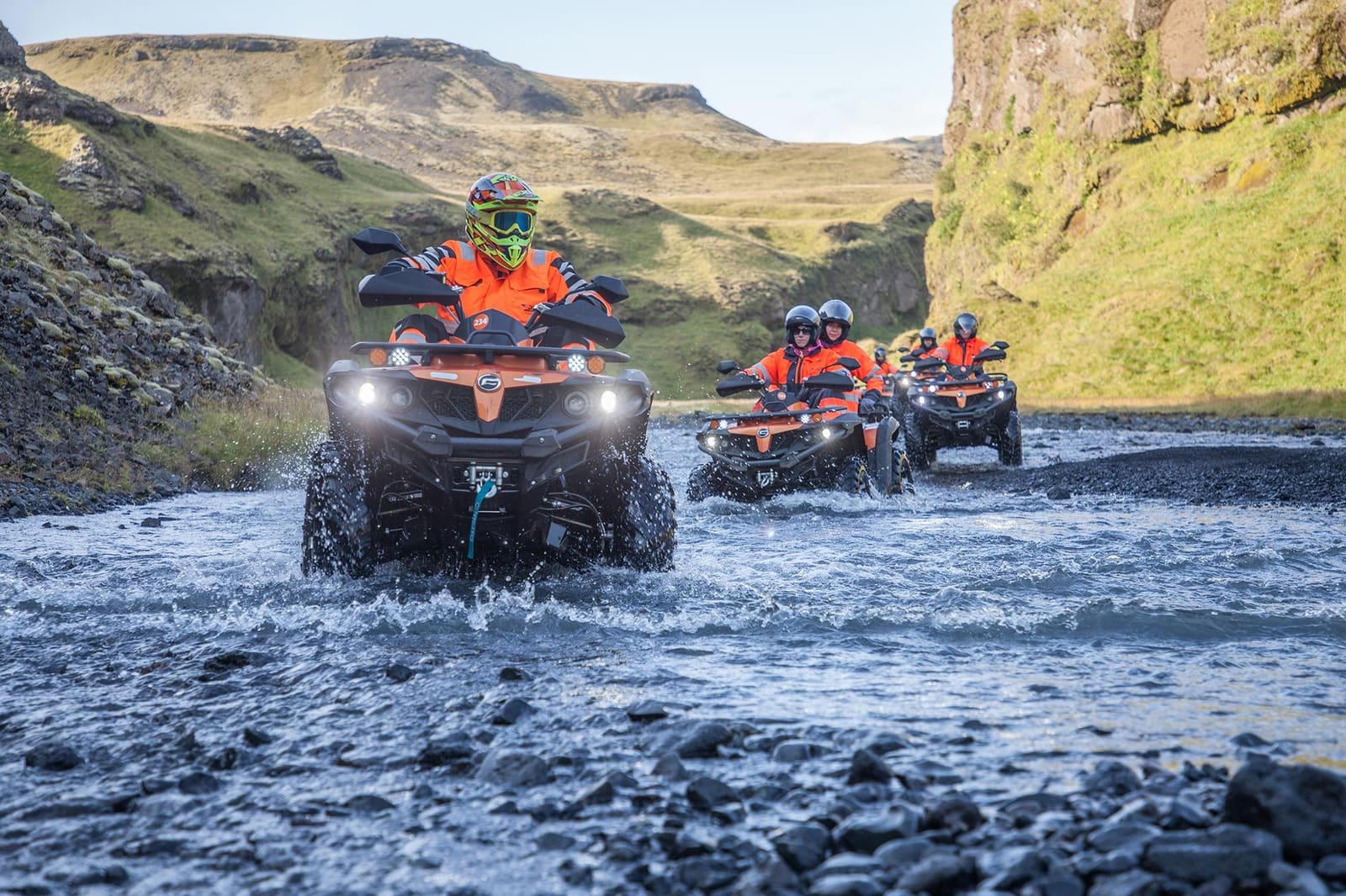 Helmeted ATV riders crossing a shallow stream in open Icelandic terrain.