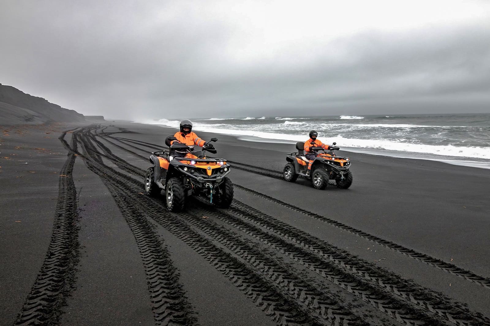 Two helmeted ATV riders following tire tracks along a South Coast black-sand beach.