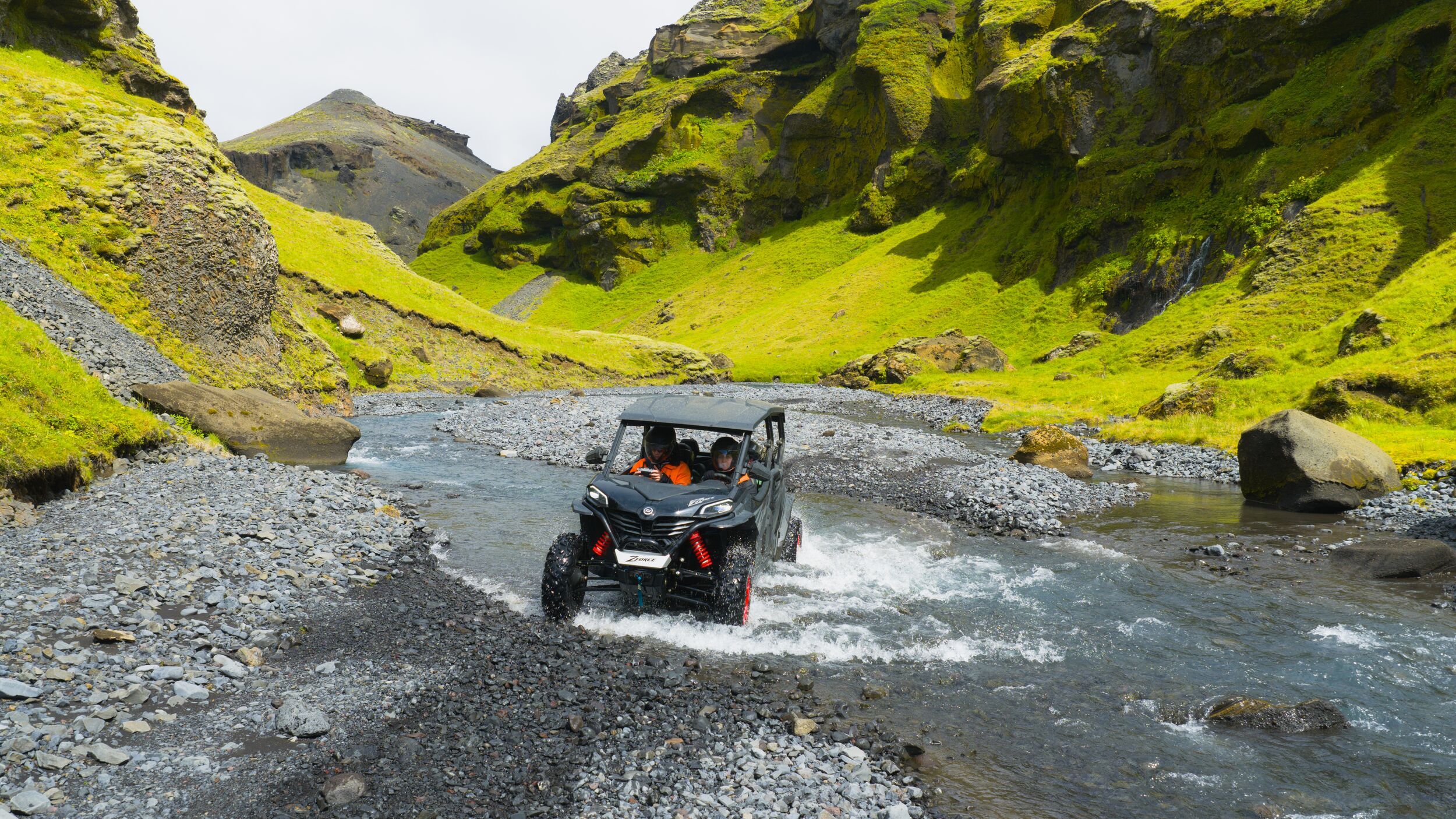 Two helmeted travelers seated side by side in a buggy crossing a shallow Icelandic stream.