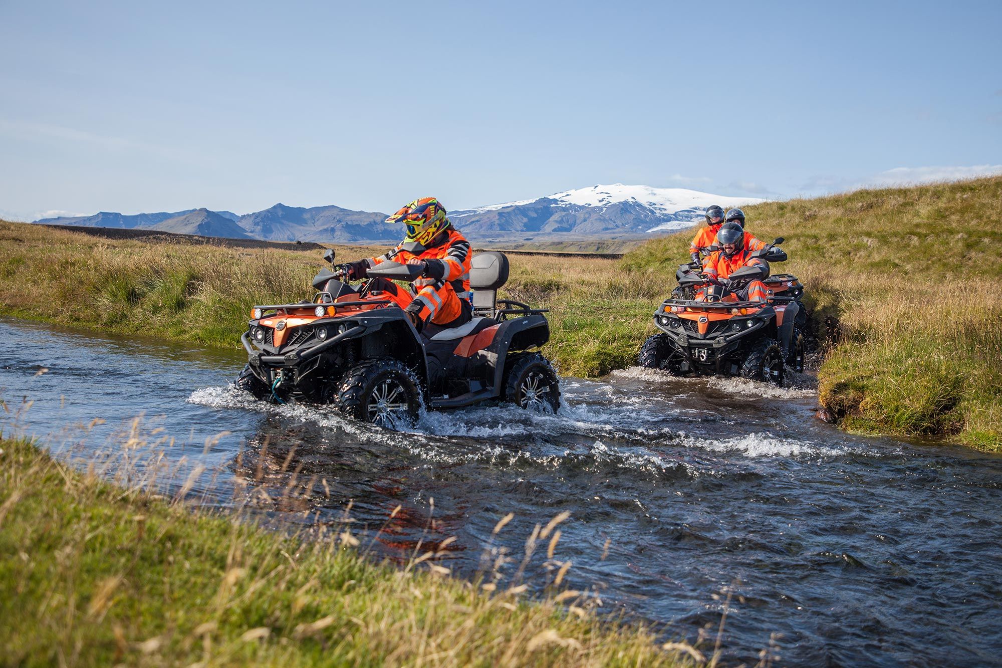 Helmeted ATV riders moving through a shallow stream below Icelandic mountains.