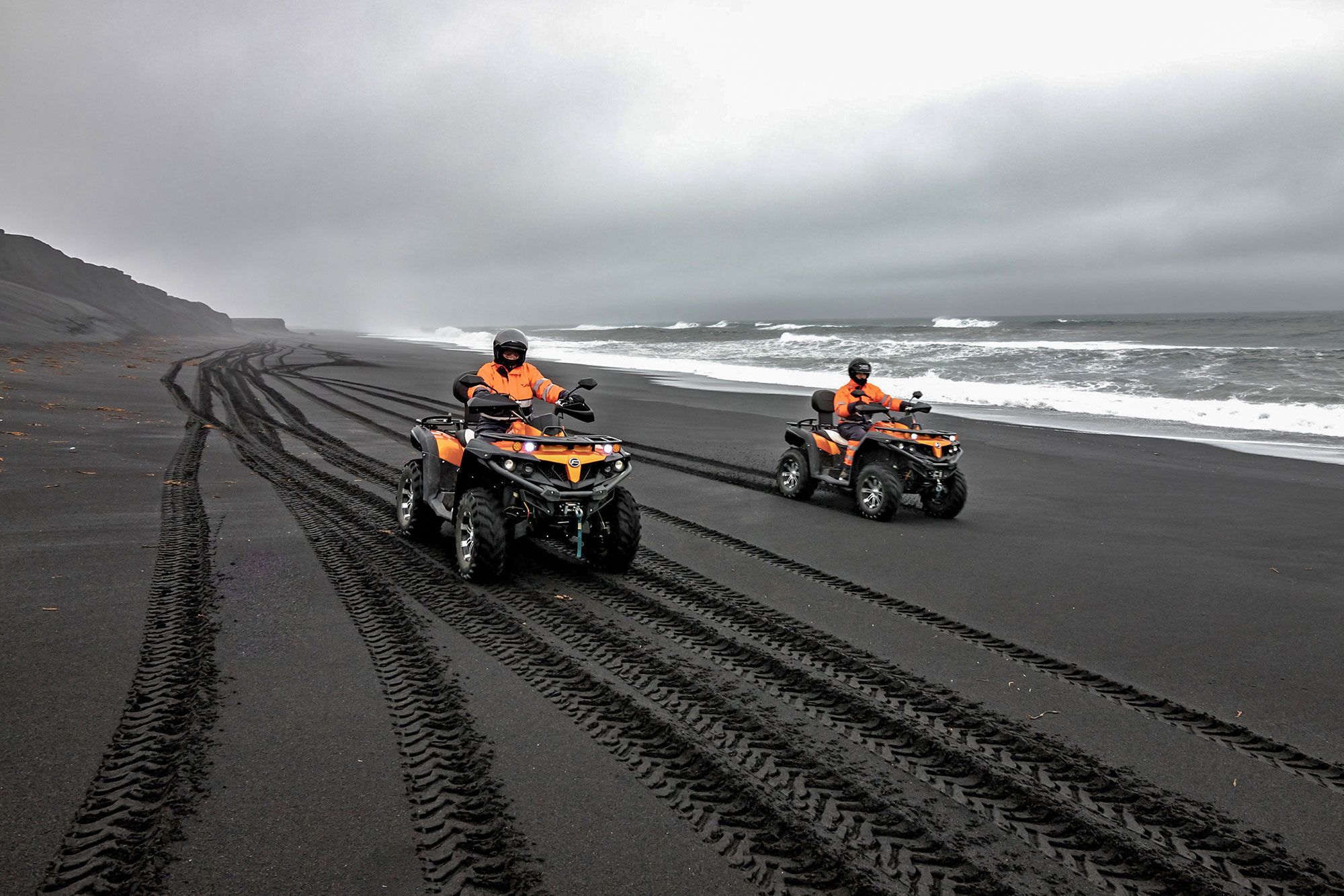 Two helmeted ATV riders following tire tracks along a South Coast black-sand beach.
