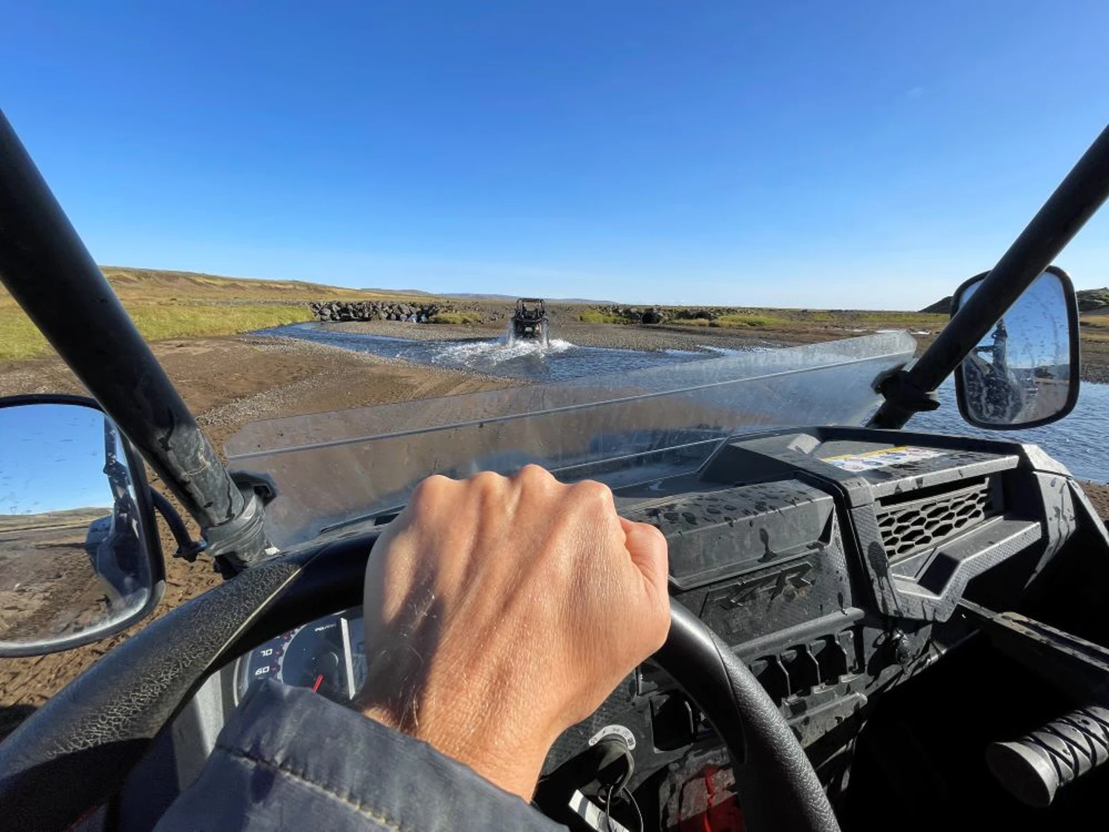 A driver's hand on a buggy steering wheel while following another buggy across a wet track.