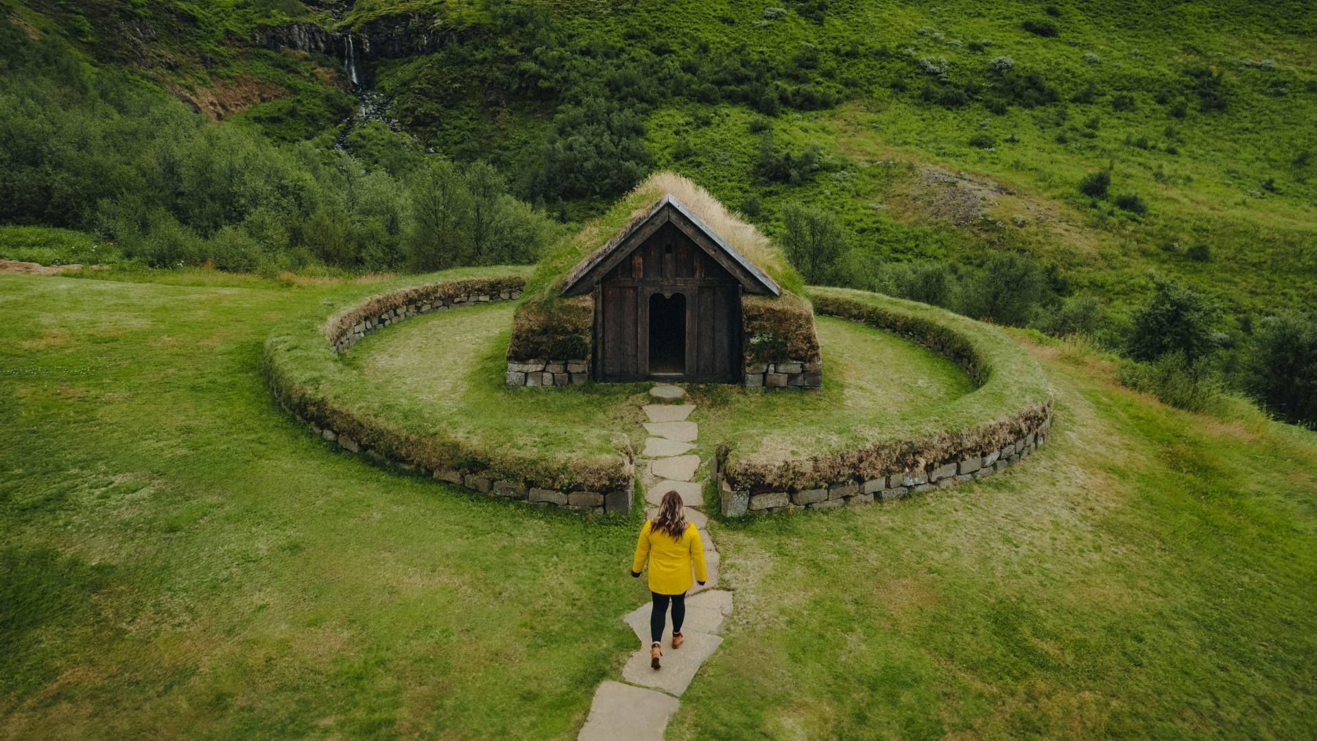 Traveler in a yellow jacket walking toward a turf-roofed Icelandic building