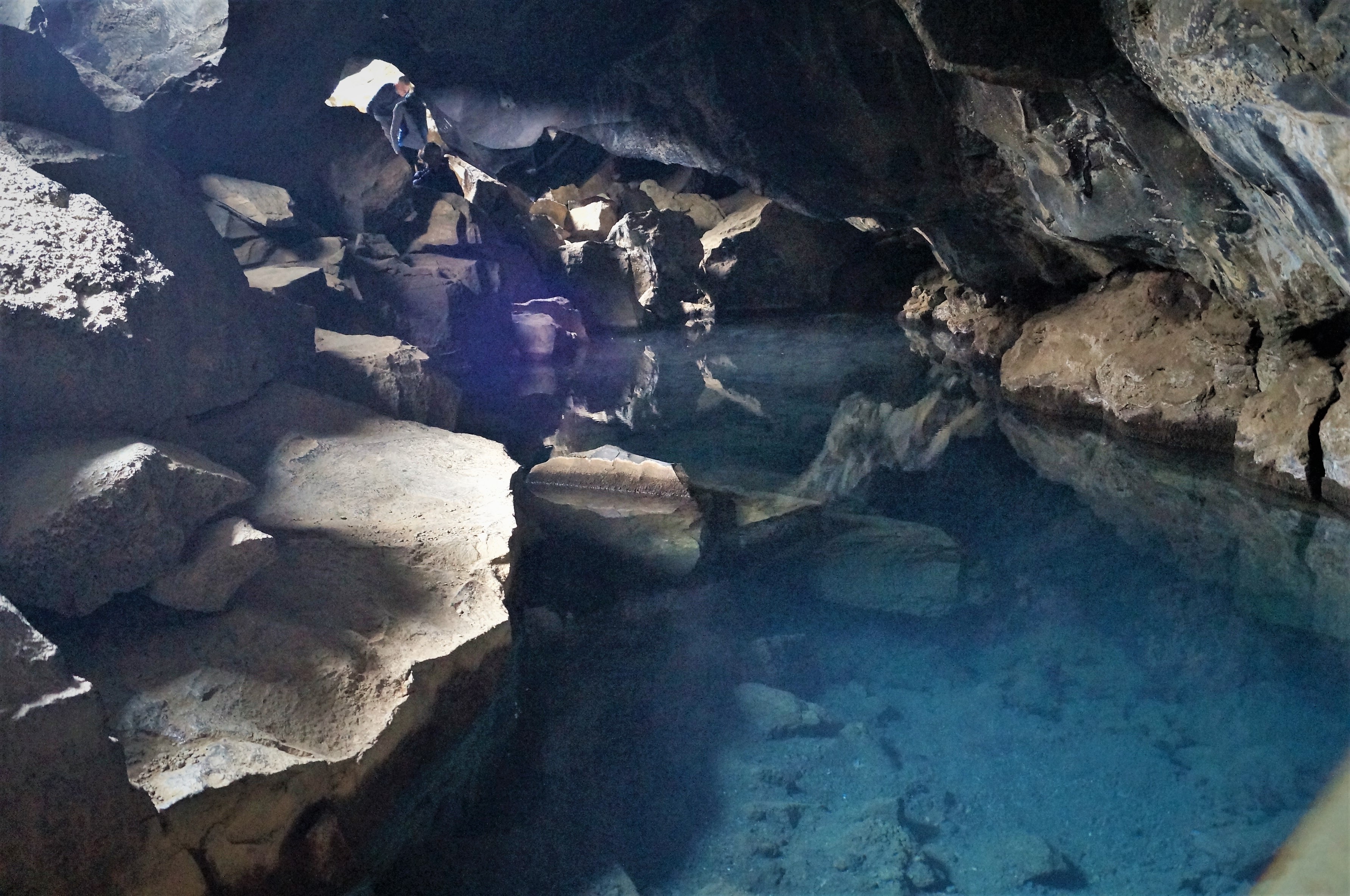 Clear pool and boulders beneath the rock ceiling of Grjótagjá, with visitors near the entrance