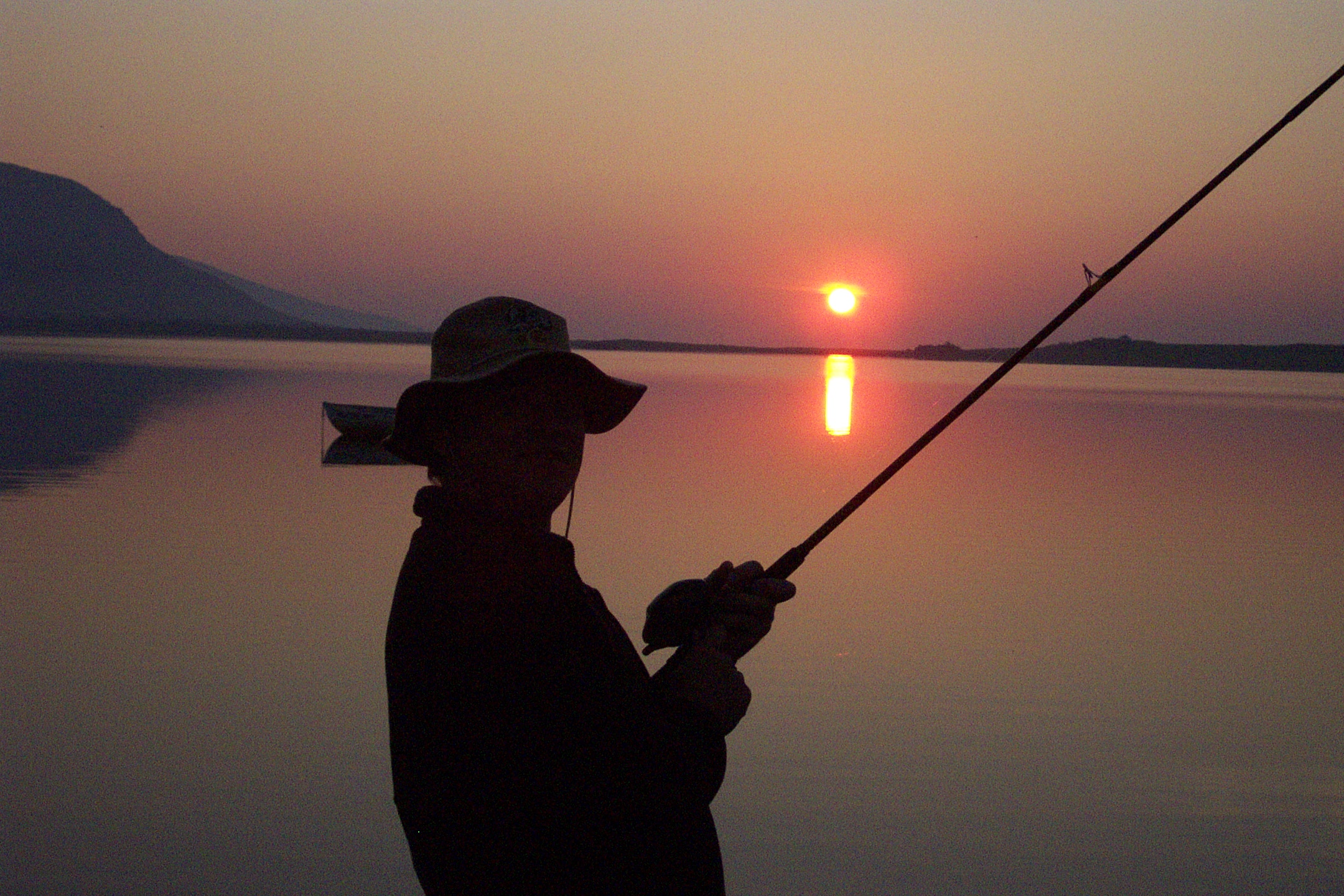 Angler holding a rod beside Meðalfellsvatn under a low orange sun