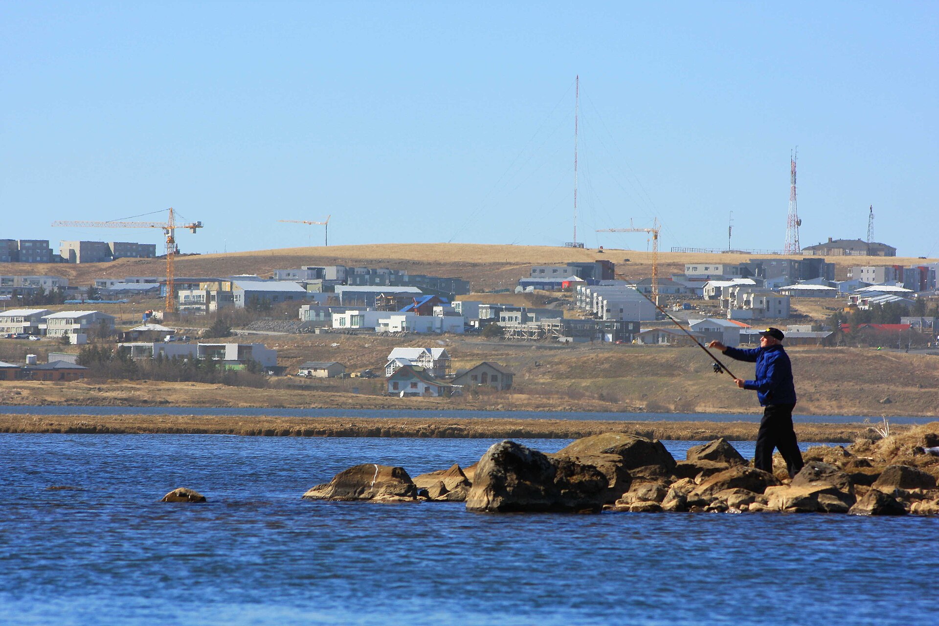 Angler casting from rocks at Elliðavatn with Reykjavík-area buildings beyond