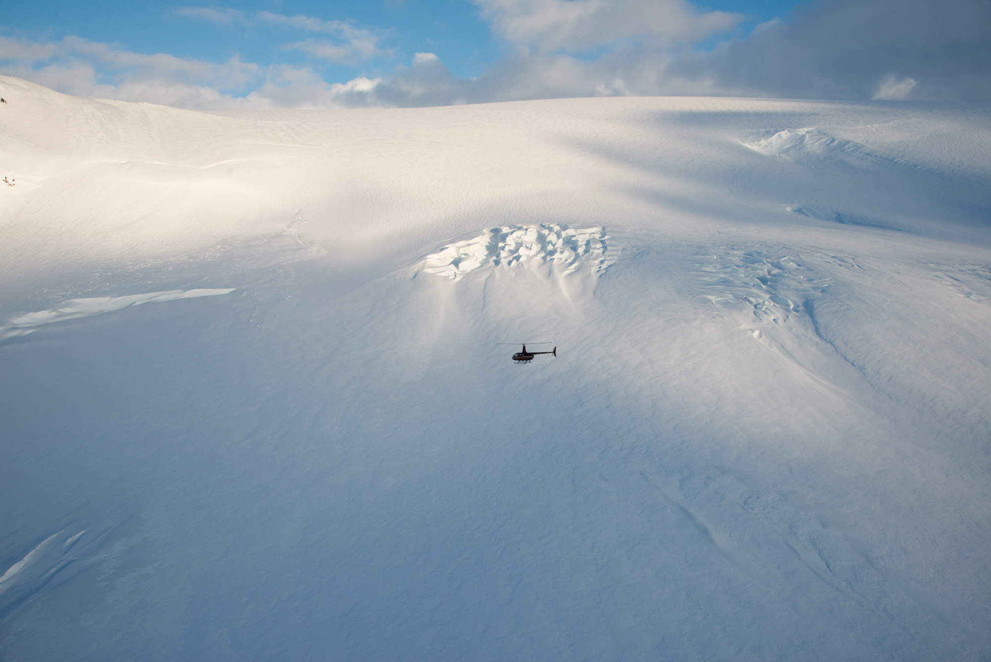 A small helicopter flying across a broad Icelandic glacier beneath broken cloud.