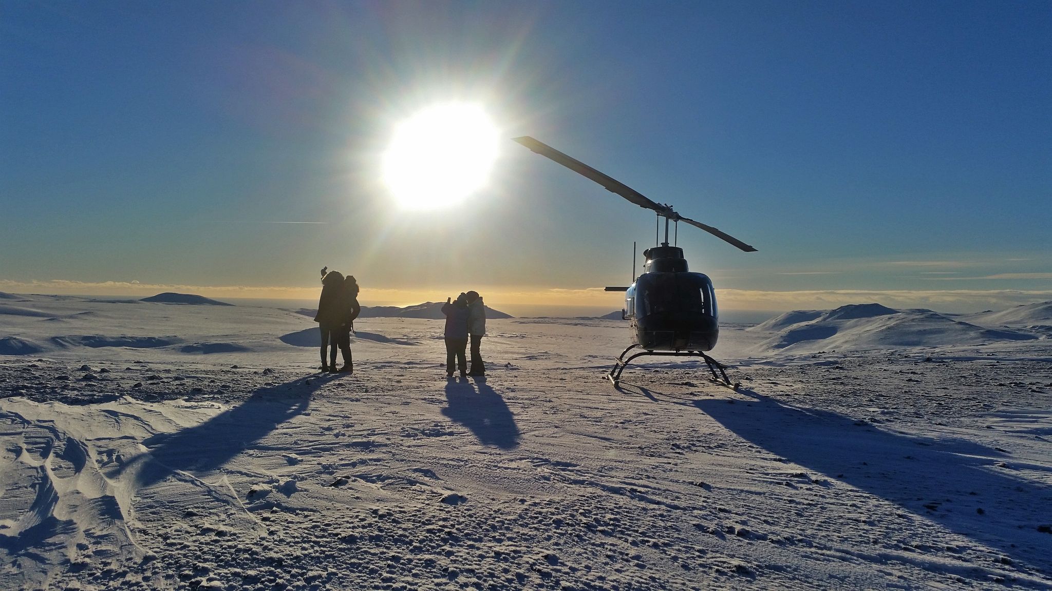 Four travelers taking photographs beside a helicopter on a snowy Icelandic landing site.