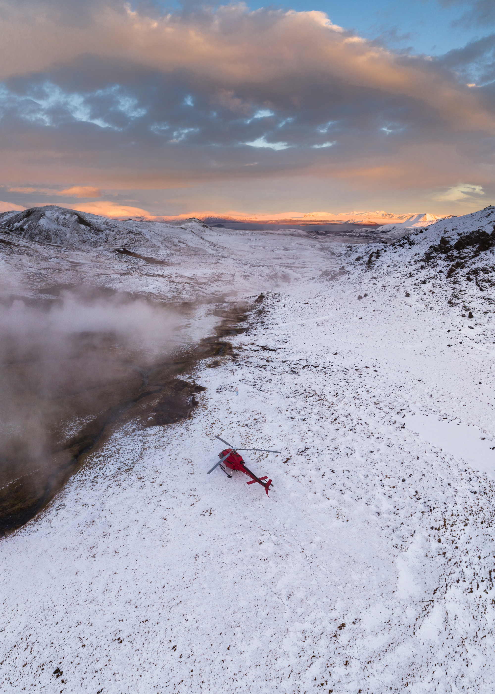 A red helicopter landed beside a steaming geothermal area in Icelandic winter conditions.
