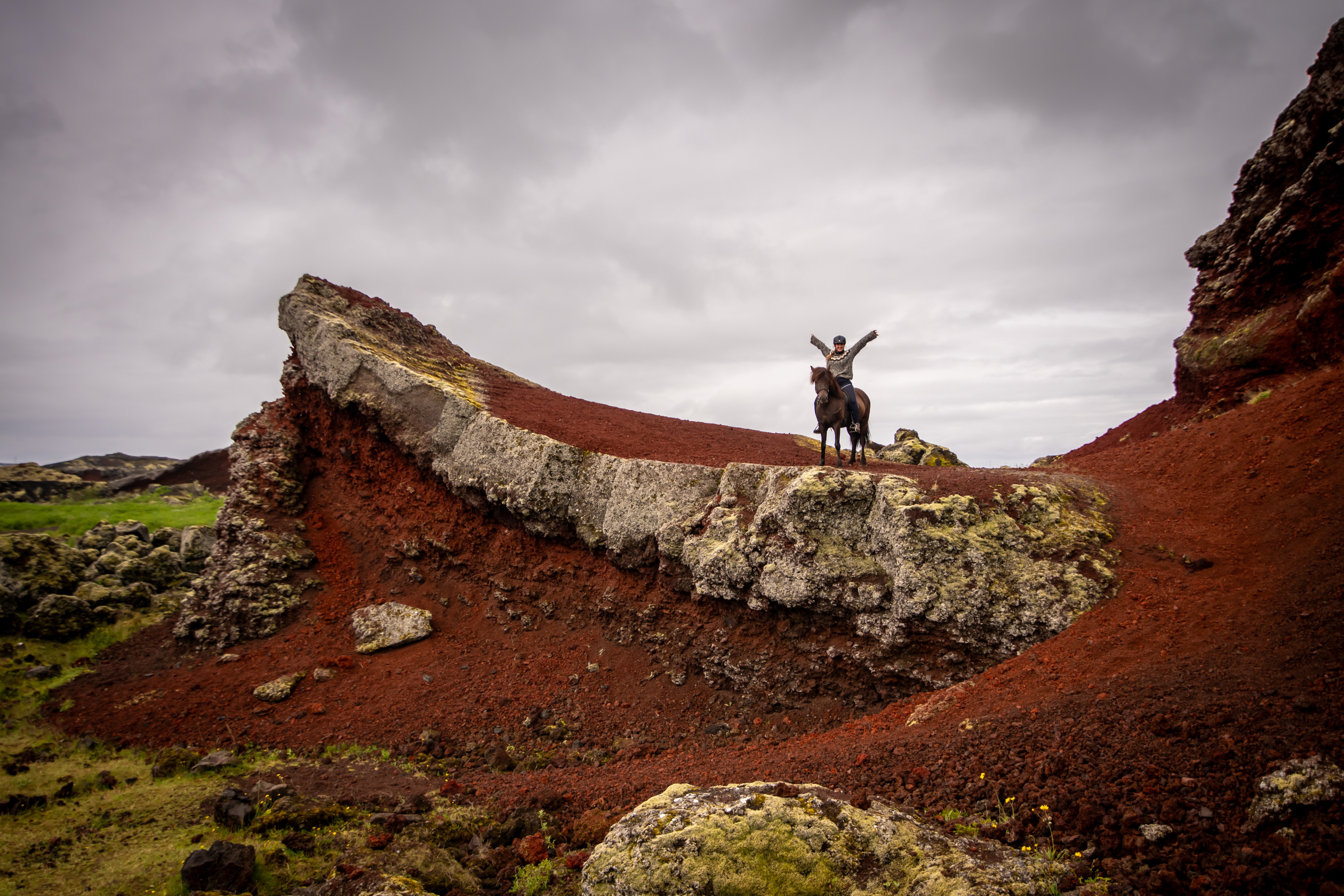 Two riders on Icelandic horses beside red volcanic rock and green moss