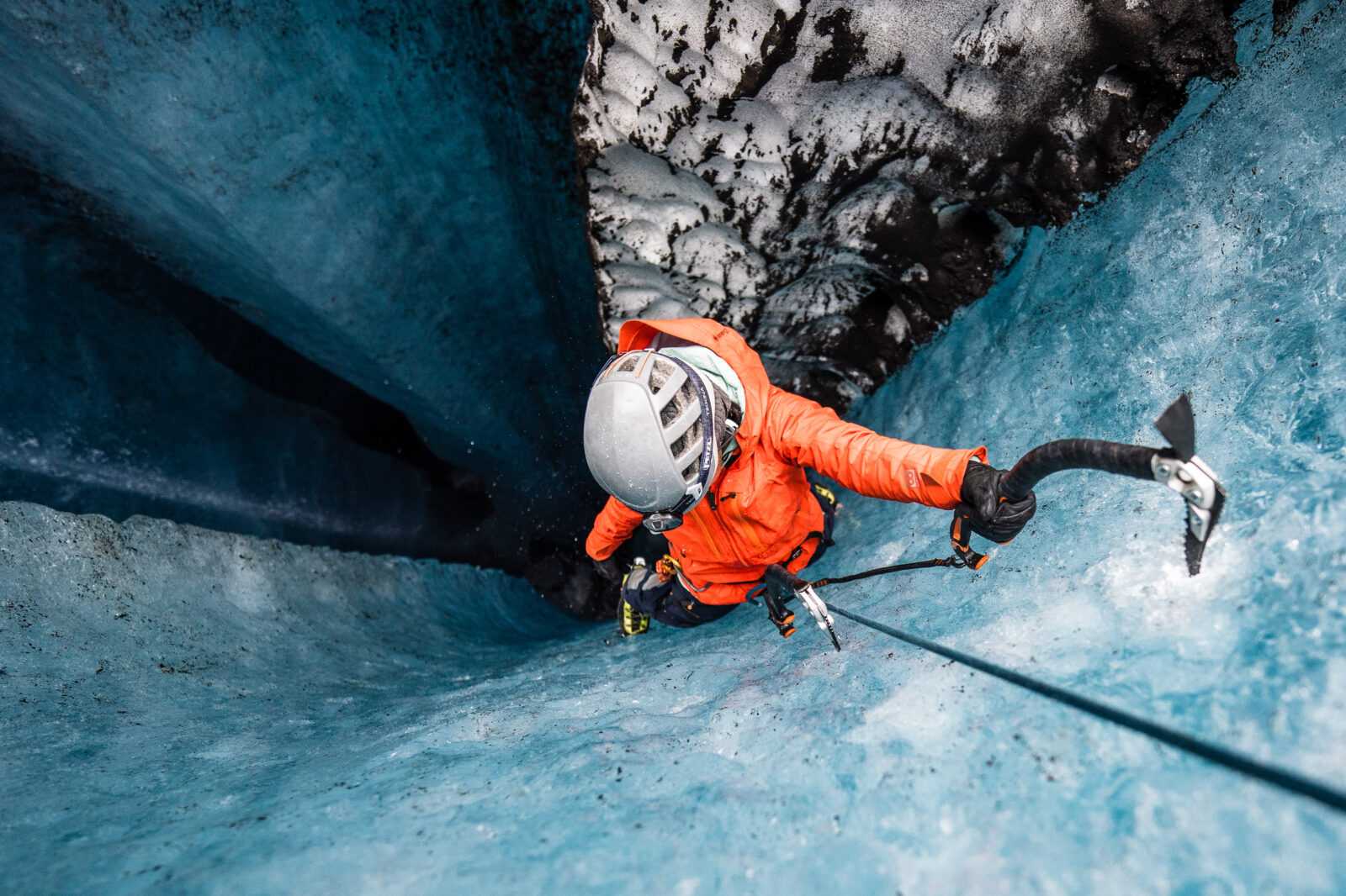 Ice climber in an orange jacket seen from above with axes, helmet, harness, and rope