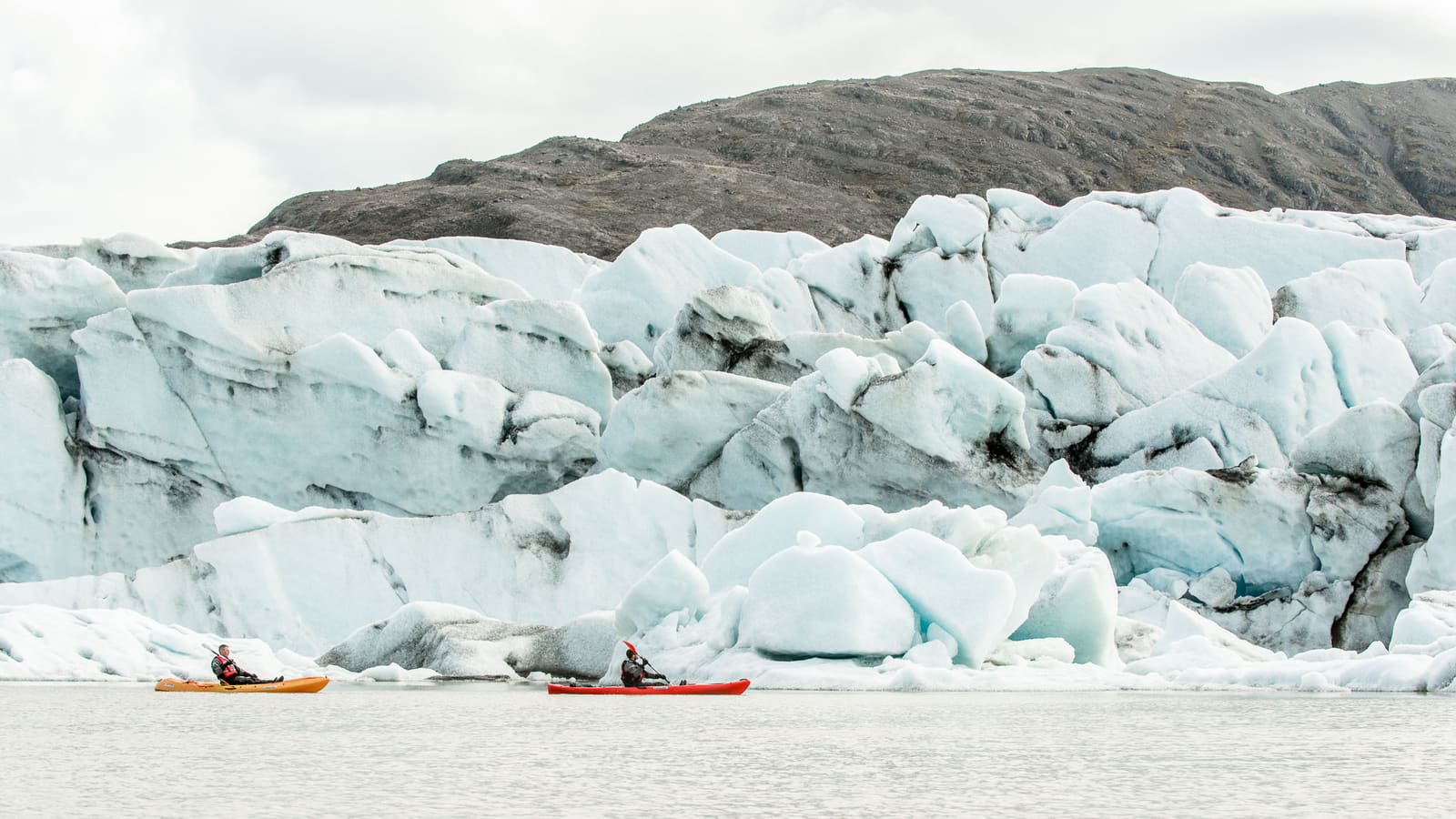 Two kayakers paddling below a wall of pale blue glacier ice in Iceland