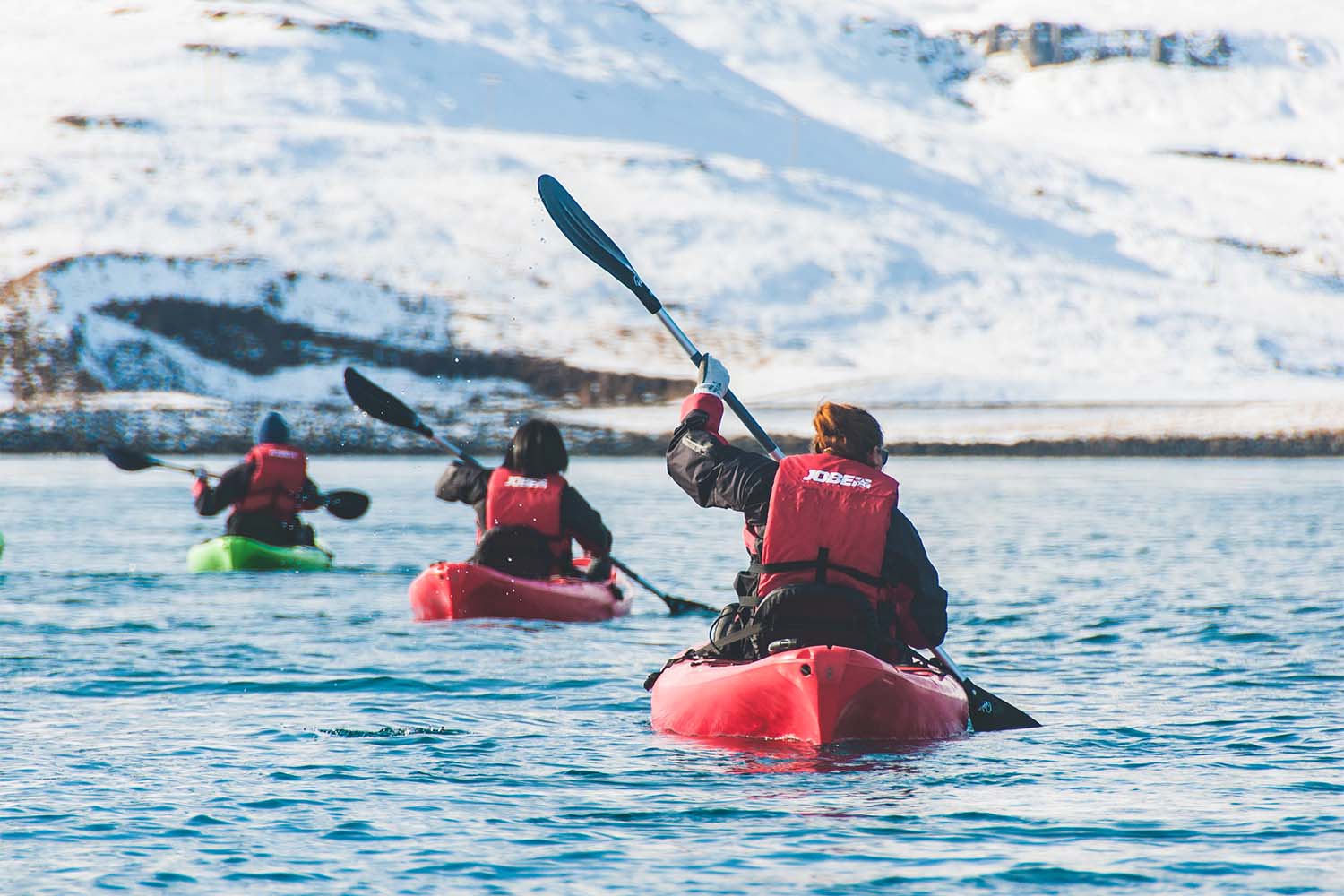 Three kayakers in cold-water gear paddling toward snowy Icelandic slopes